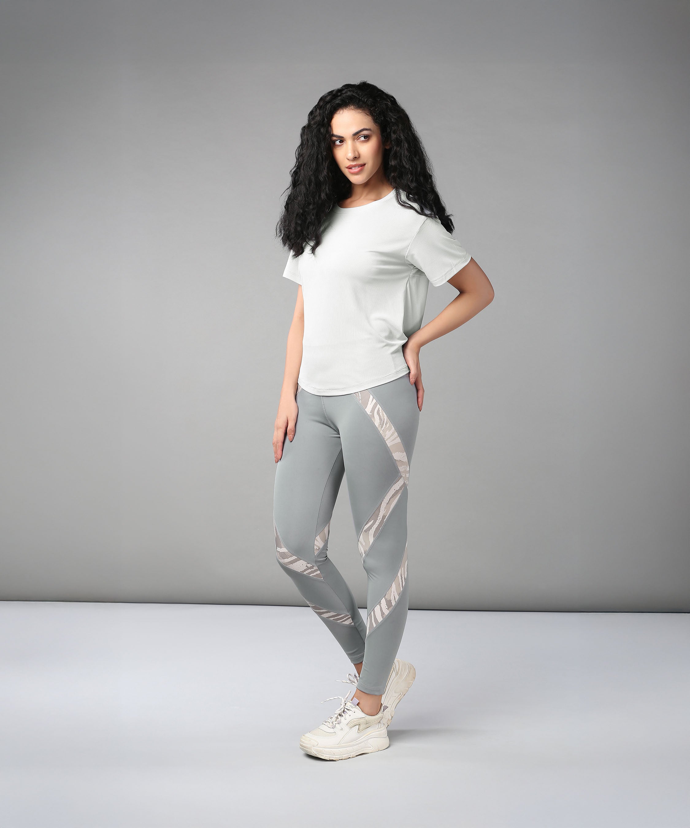 A woman showcases the Grey Top with Patched Legging, posing in a studio with a neutral gray background to highlight its super soft fabric and modern cross body design.