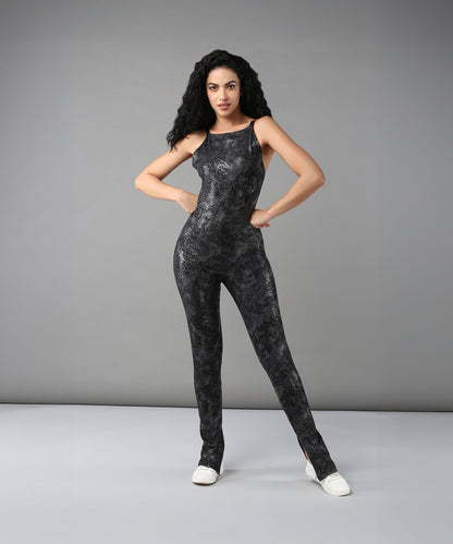 A woman with long curly hair poses in the Glistening Nightfall Jump Suit, featuring a black patterned polyblend design, paired with white shoes against a gray background.