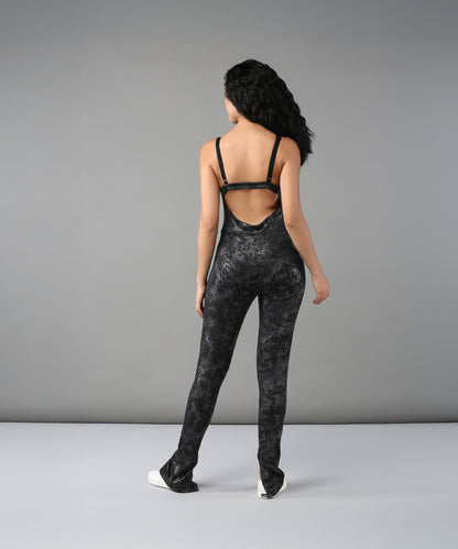 Woman with long hair wears the Glistening Nightfall Jump Suit—a black, open-back polyblend jumpsuit with adjustable straps—paired with white shoes, standing against a gray background.
