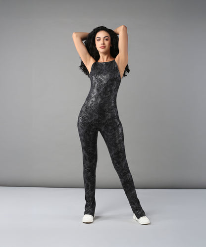 A woman poses with hands behind her head against a gray background, wearing the Glistening Nightfall Jump Suit featuring a black pattern and paired with white shoes.