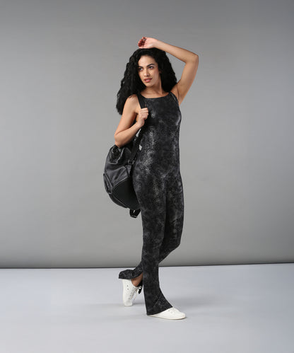 A woman wearing the Glistening Nightfall Jump Suit—black with a snakeskin pattern—poses against a gray background, styled with white sneakers and a backpack.
