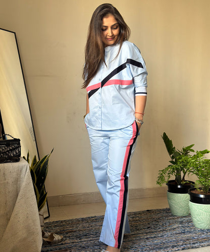 A woman wears the Contrast Cross Batwing Shirt With Wide Legged Bottom set in blue with pink and black stripes, standing indoors near plants, looking down and smiling.