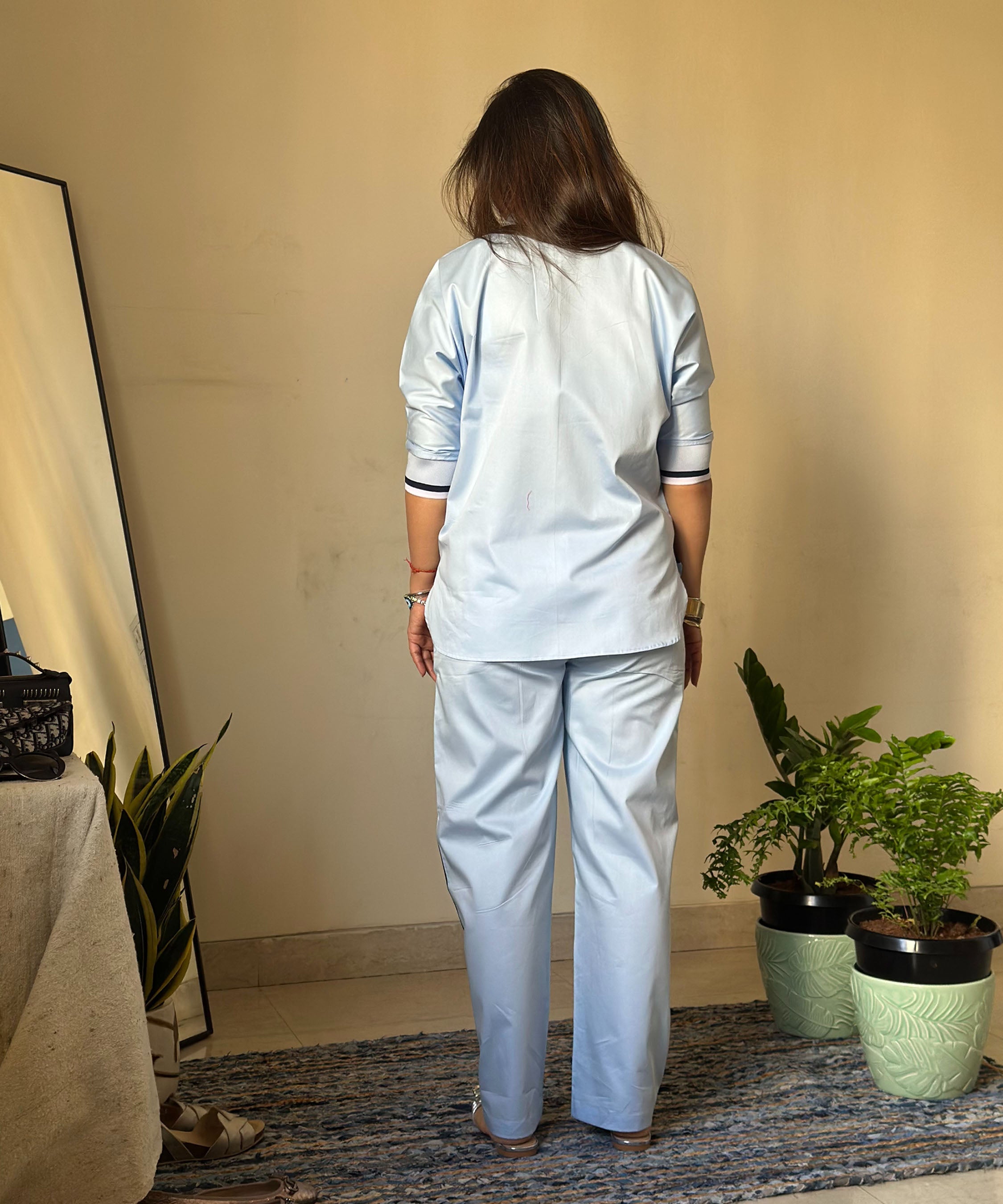 A woman wearing the Contrast Cross Batwing Shirt With Wide Legged Bottom stands facing away beside potted plants and a mirror in a minimalist room.