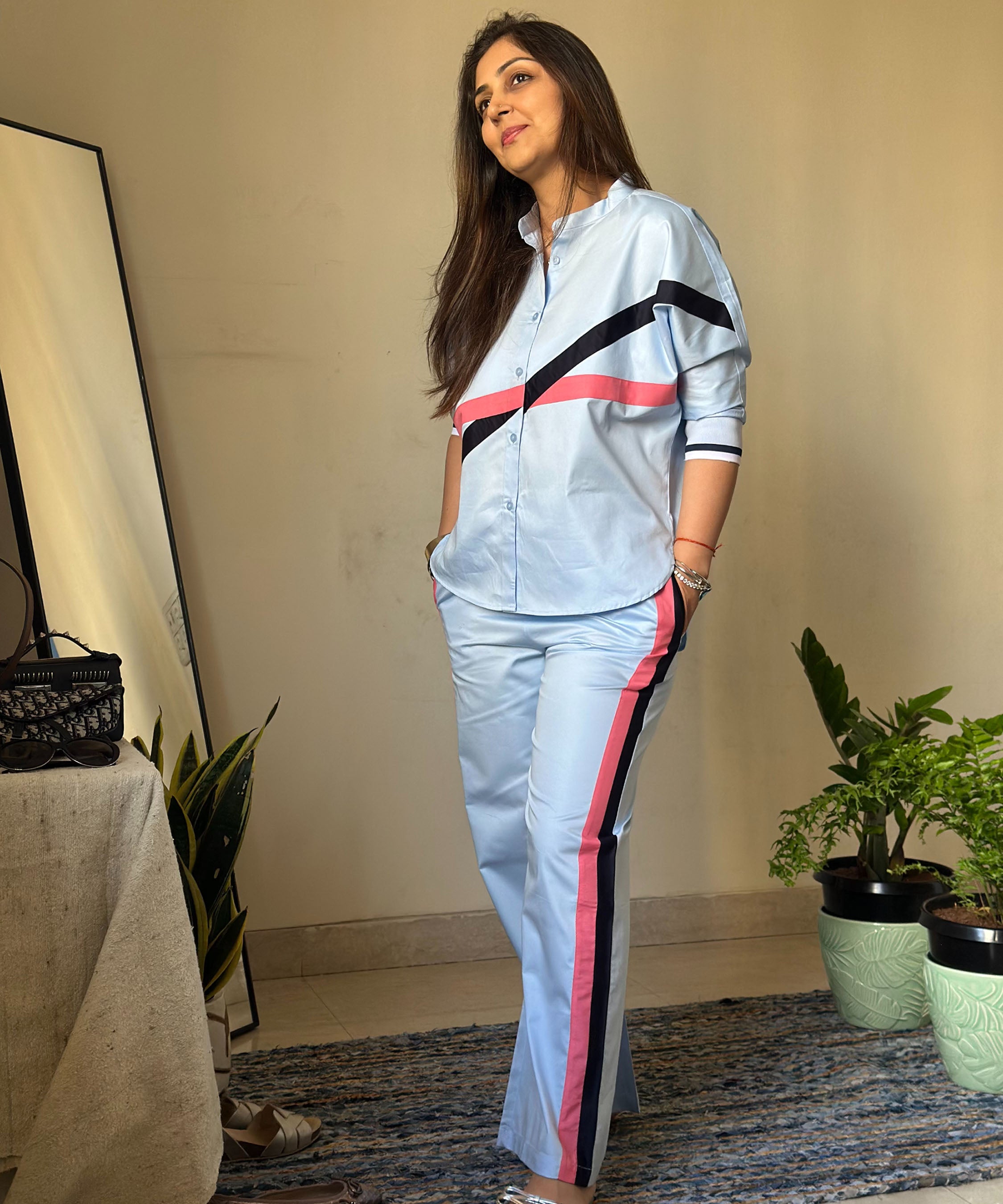 A woman stands near plants and a mirror in a cozy room, wearing the Contrast Cross Batwing Shirt with Wide Legged Bottom—a blue joggers set featuring black and pink stripes and comfy batwing sleeves.