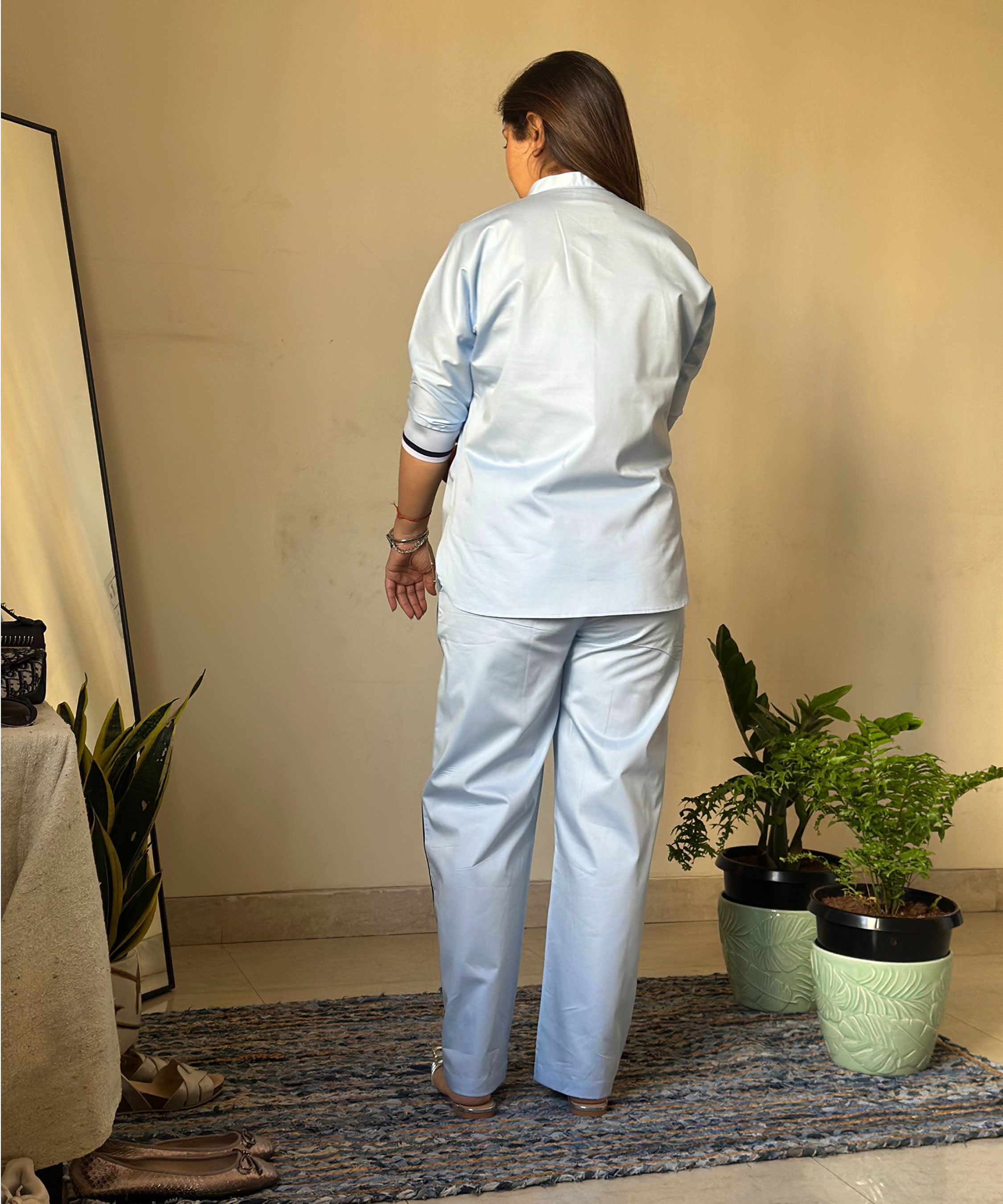 A woman wears the Contrast Cross Batwing Shirt With Wide Legged Bottom, standing on a rug facing away near plants and a mirror in a room.