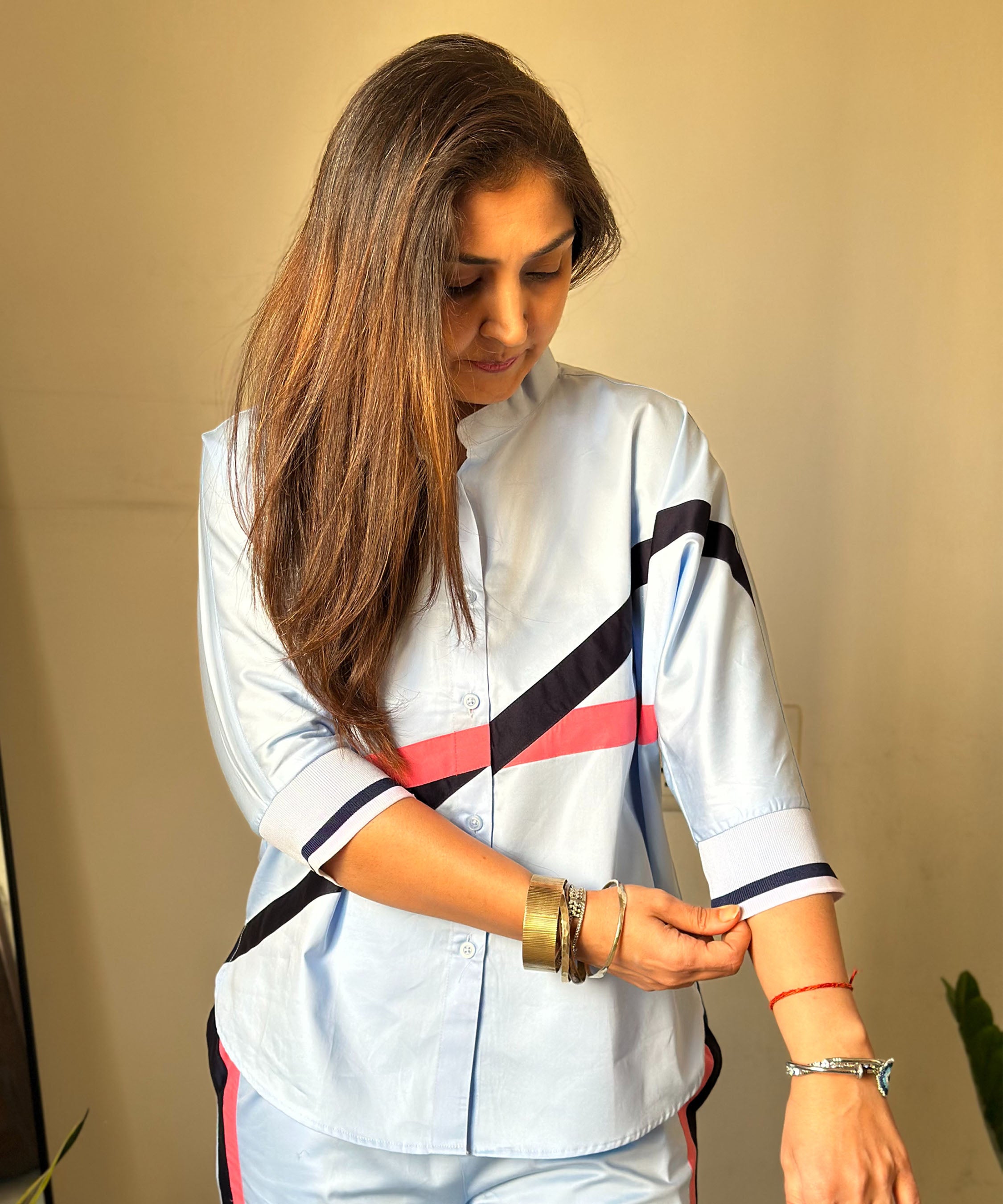 A woman in a Contrast Cross Batwing Shirt With Wide Legged Bottom, made of light blue 100% cotton, rolls up her batwing sleeve and looks down, showing bracelets and a red thread on her wrist.