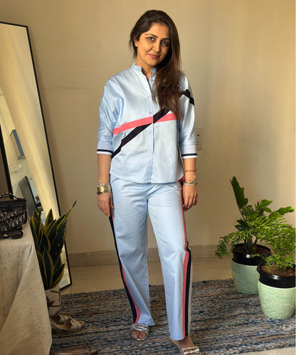 Woman wearing the Contrast Cross Batwing Shirt With Wide Legged Bottom, a 100% cotton jogger set with light blue wide-leg pants and a striped batwing sleeve shirt, standing indoors near potted plants.