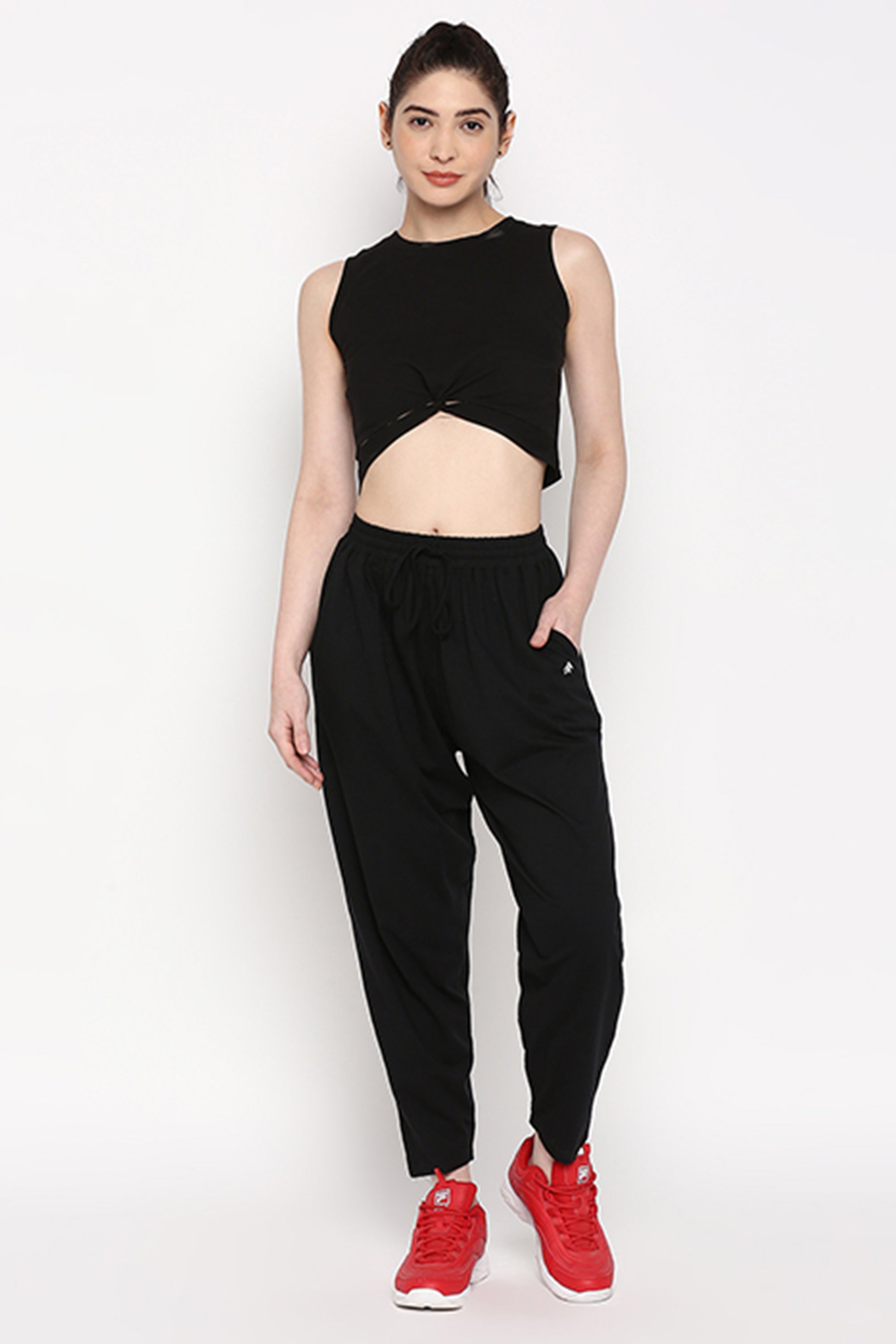 A woman wears stretchable black Yoga Pants and a black sleeveless crop top with red sneakers, posing against a white background.