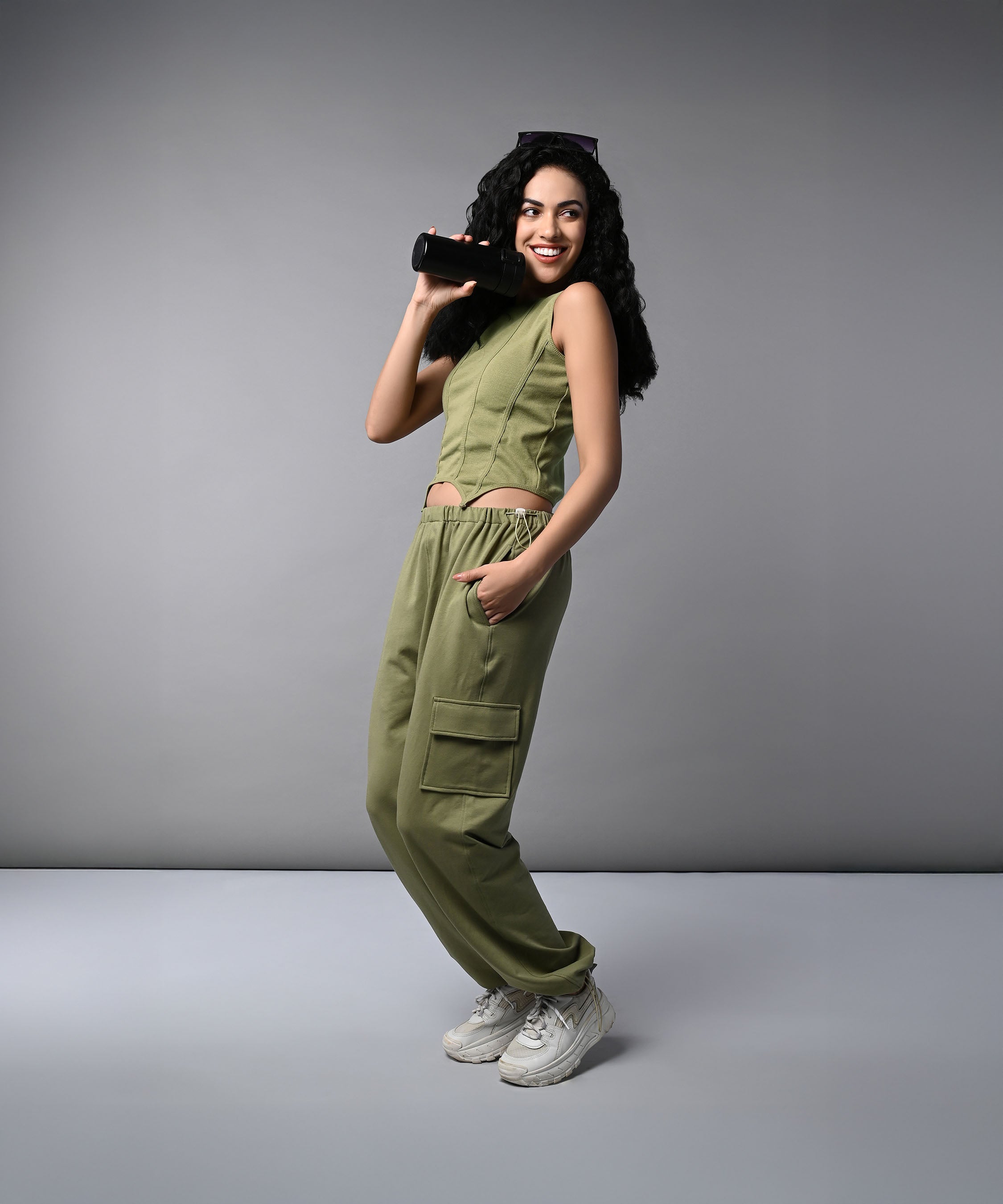 A woman in the Olive Corset Crop Top with Parachute Pants and white sneakers smiles while holding a portable speaker against a gray background.