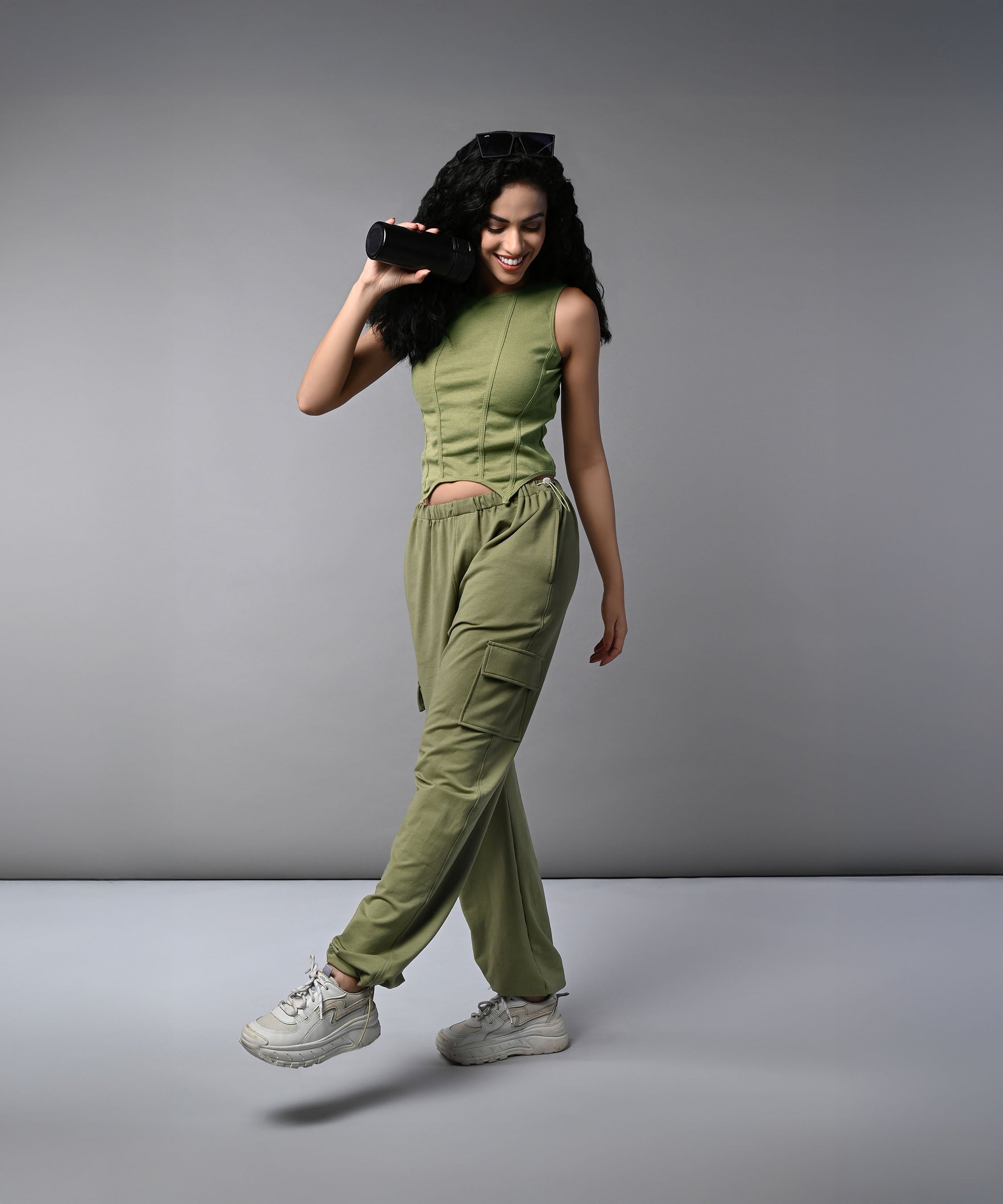 A woman smiles and walks against a plain gray background, wearing the Olive Corset Crop - Top with Parachute Pants, while holding a portable speaker.