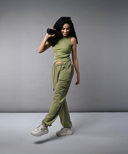 A woman smiles and walks against a plain gray background, wearing the Olive Corset Crop - Top with Parachute Pants, while holding a portable speaker.