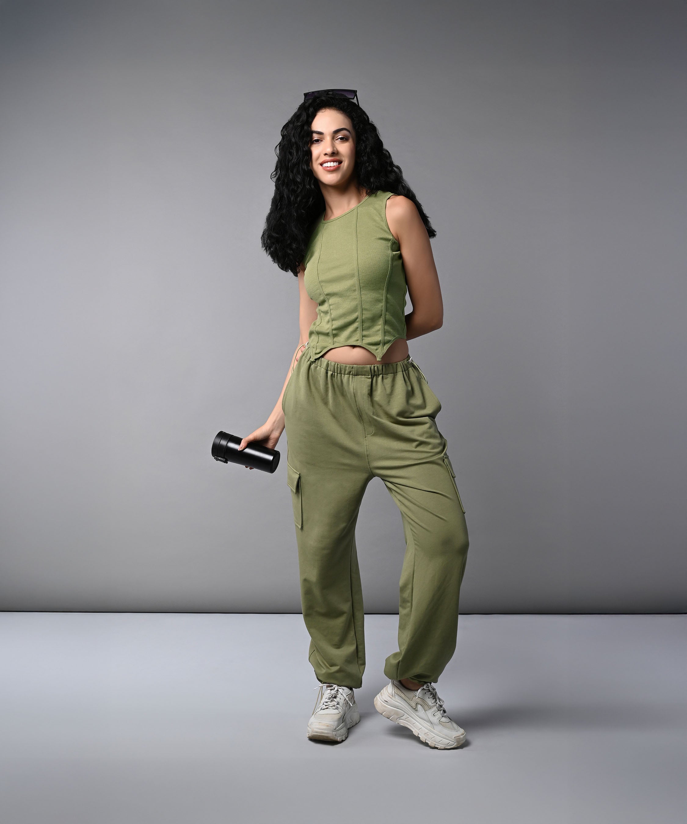 Woman wearing the Olive Corset Crop - Top with Parachute Pants, holding a water bottle and smiling against a plain grey background.