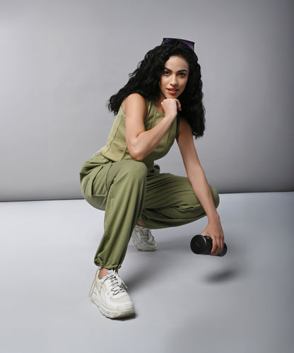 Woman wears the Olive Corset Crop - Top with Parachute Pants co-ord, crouching and holding a dumbbell against a plain gray background.