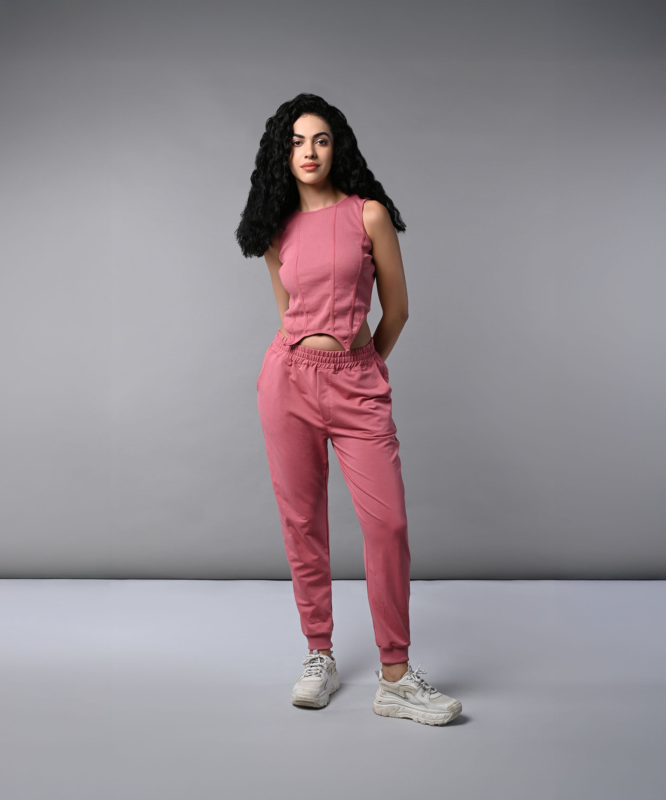 A woman with long curly hair wears a Pink Corset Crop-Top with Joggers and white sneakers, standing against a plain gray background.