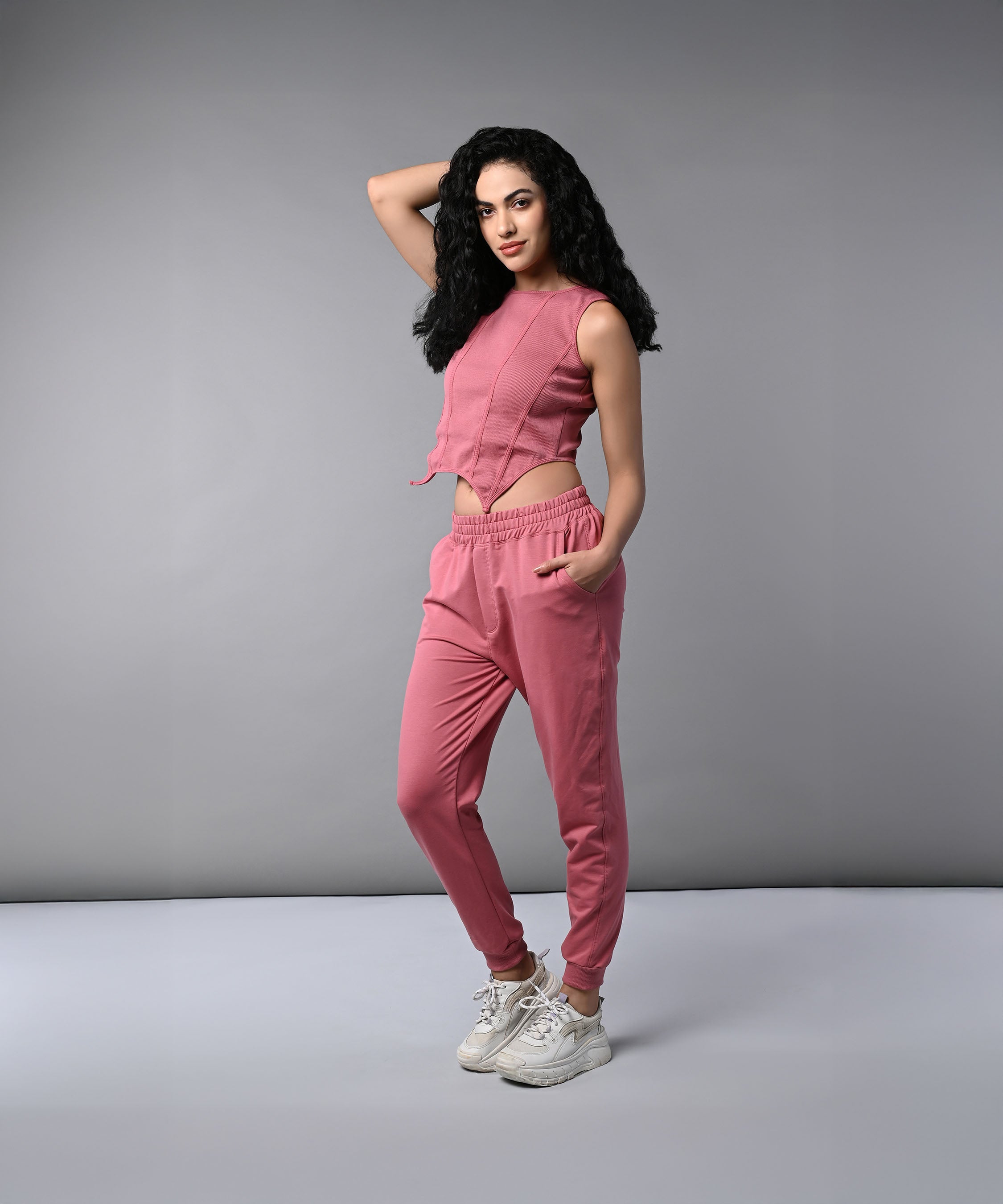 A woman confidently poses against a plain gray background wearing a Pink Corset Crop-Top with Joggers.