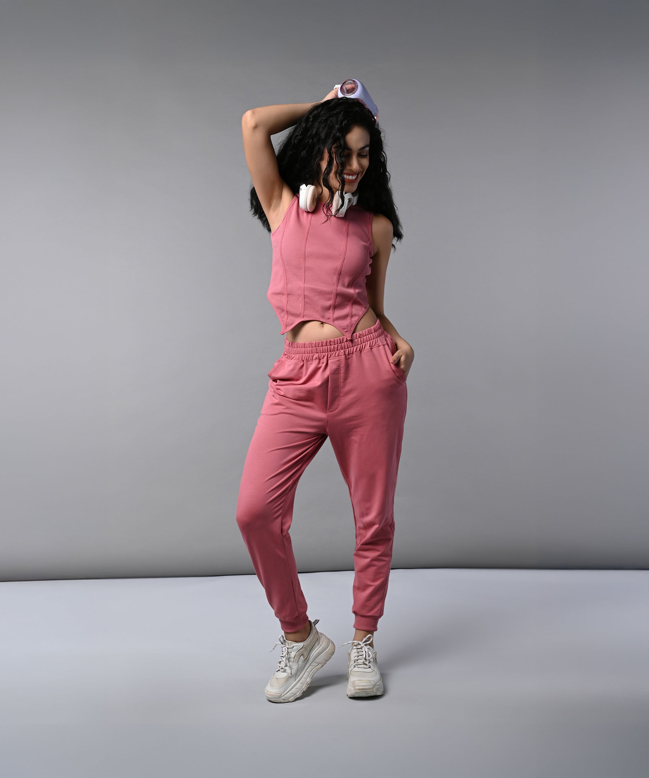 Woman wears a Pink Corset Crop-Top with Joggers, paired with headphones, posing confidently against a gray background.