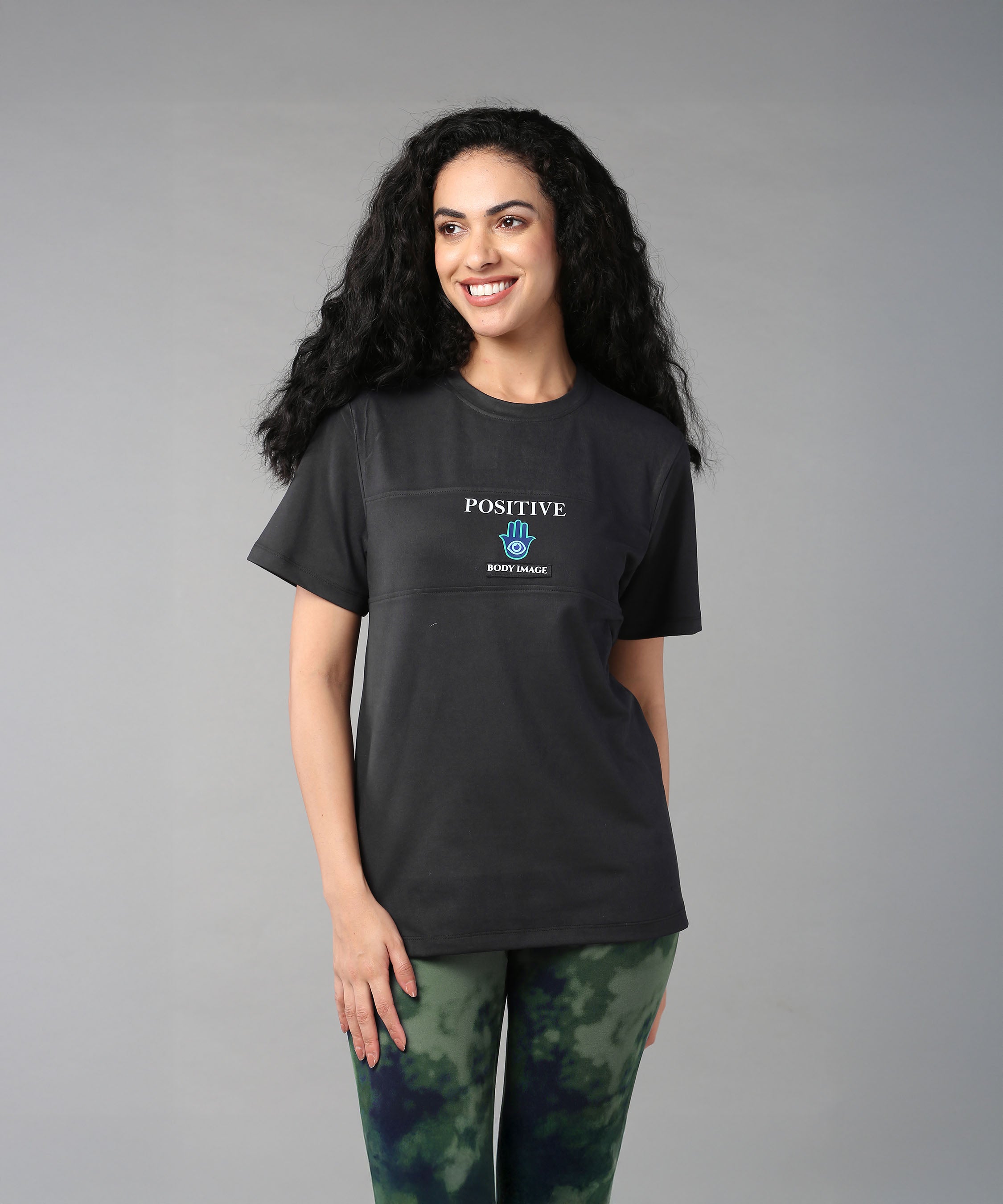A smiling woman with long curly hair wears the Positive Body Image T Shirt in black, paired with tie-dye leggings.