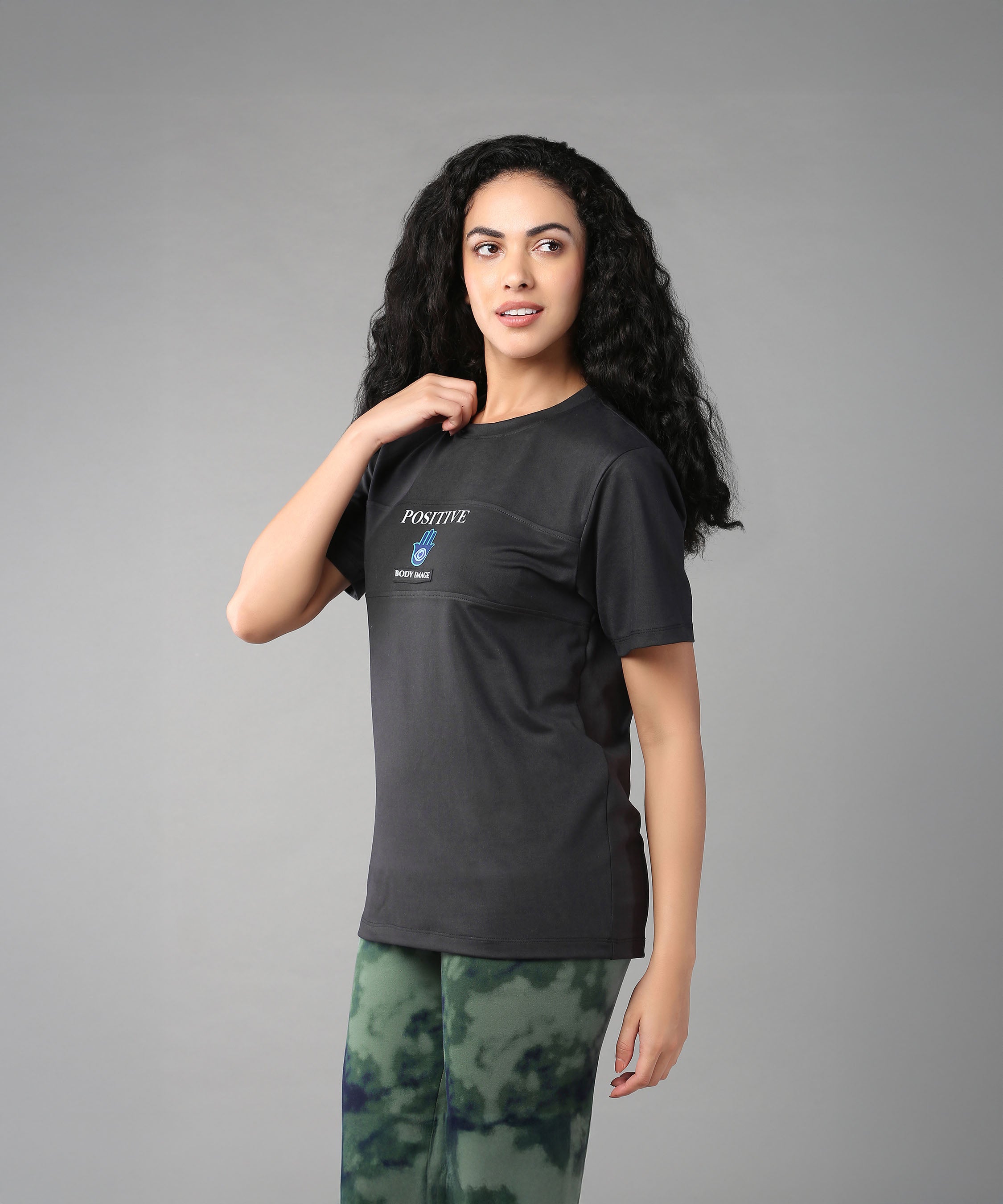 A woman with long curly hair embraces body positivity in the Positive Body Image T Shirt, featuring "Positive" text, styled with green tie-dye pants against a gray background.