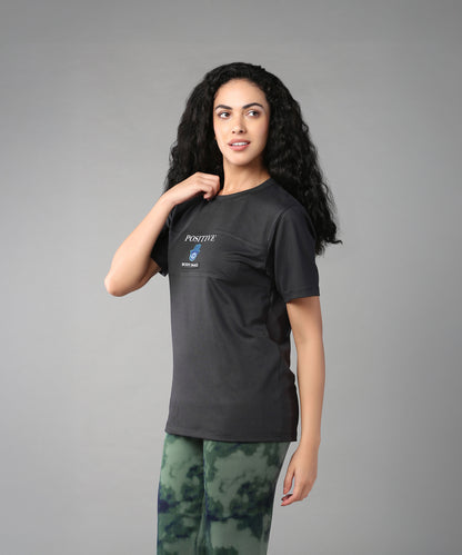 A woman with long curly hair embraces body positivity in the Positive Body Image T Shirt, featuring "Positive" text, styled with green tie-dye pants against a gray background.