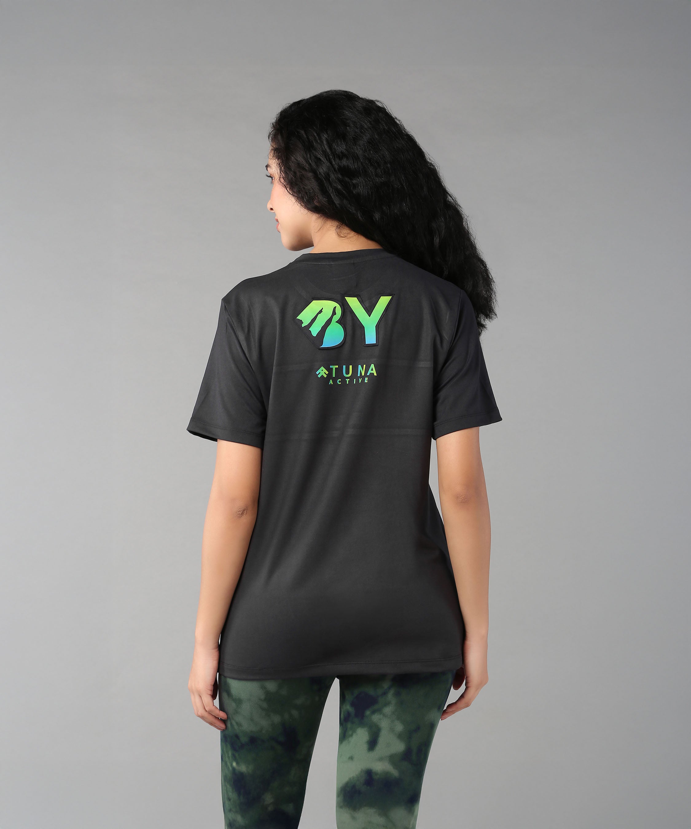 A woman with long curly hair, seen from behind, wears the Positive Body Image T Shirt—a black basic tee with green text and half sleeves—embracing positive body image.