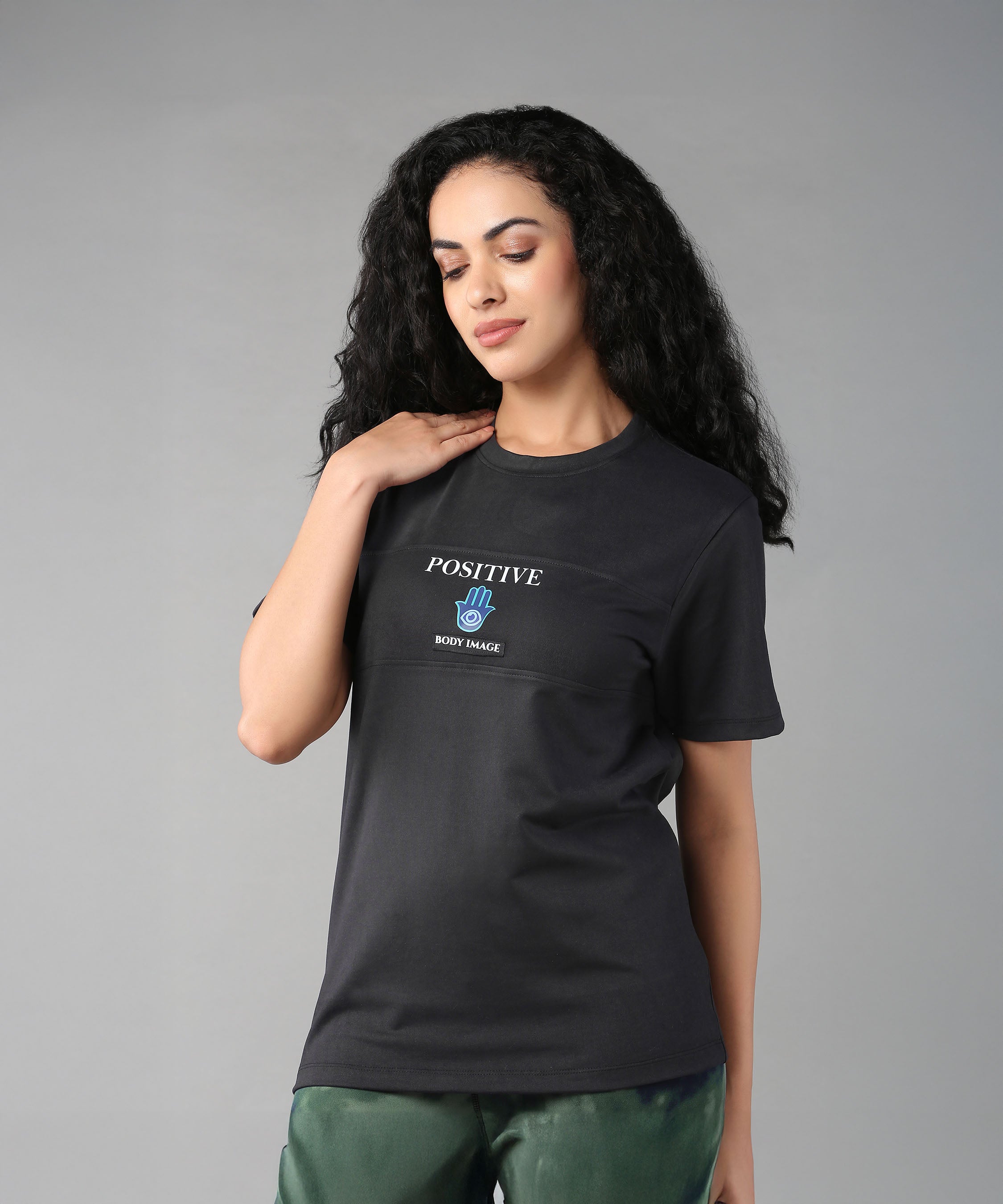 A woman with long curly hair stands against a gray background, wearing the Positive Body Image T Shirt, a black half-sleeve basic tee with "Positive Body Image" printed on it, highlighting her confident and empowering presence.