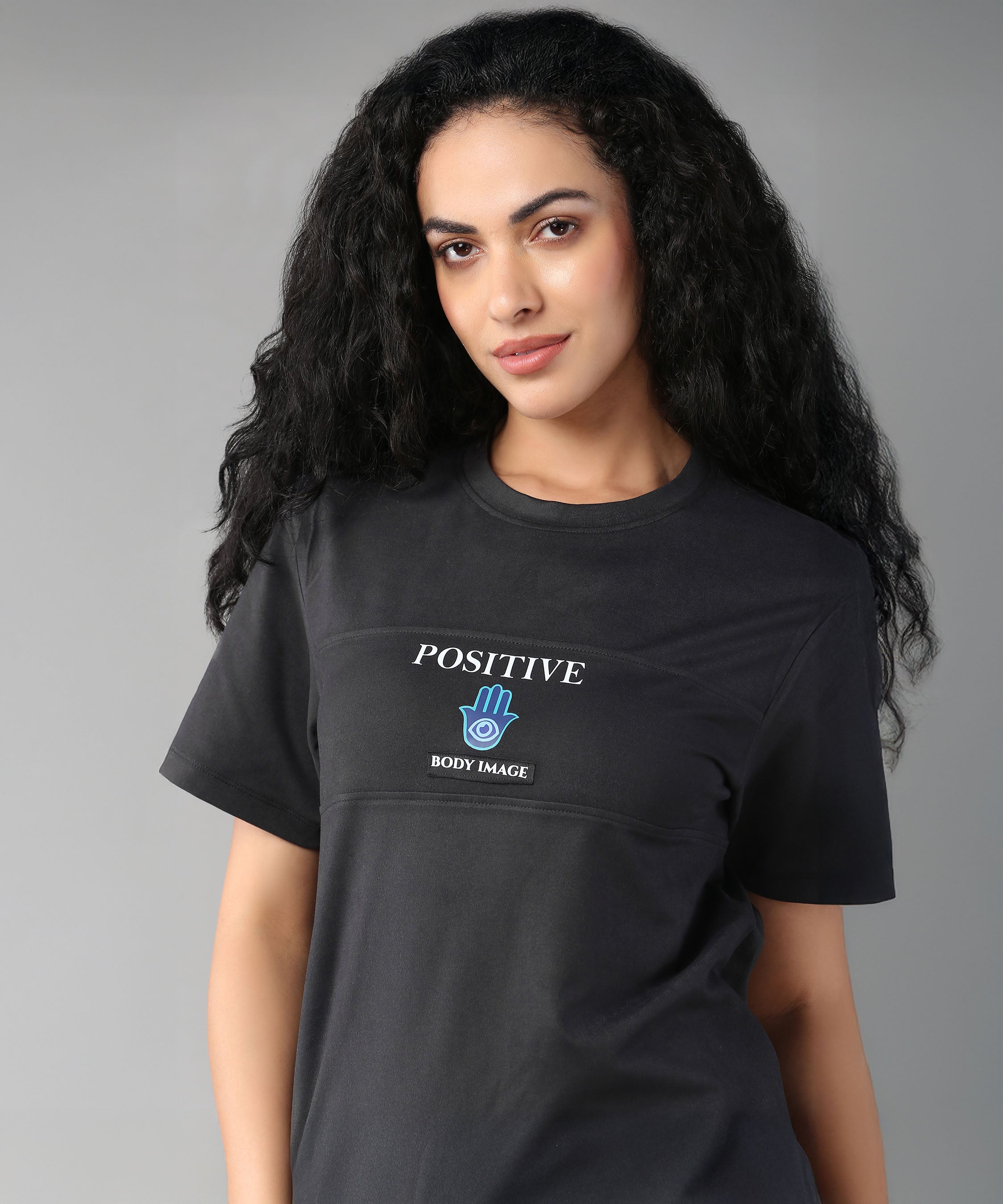 A woman with long curly hair smiles against a gray background, wearing the Positive Body Image T Shirt—a black, basic half-sleeve tee with a "Positive Body Image" print.