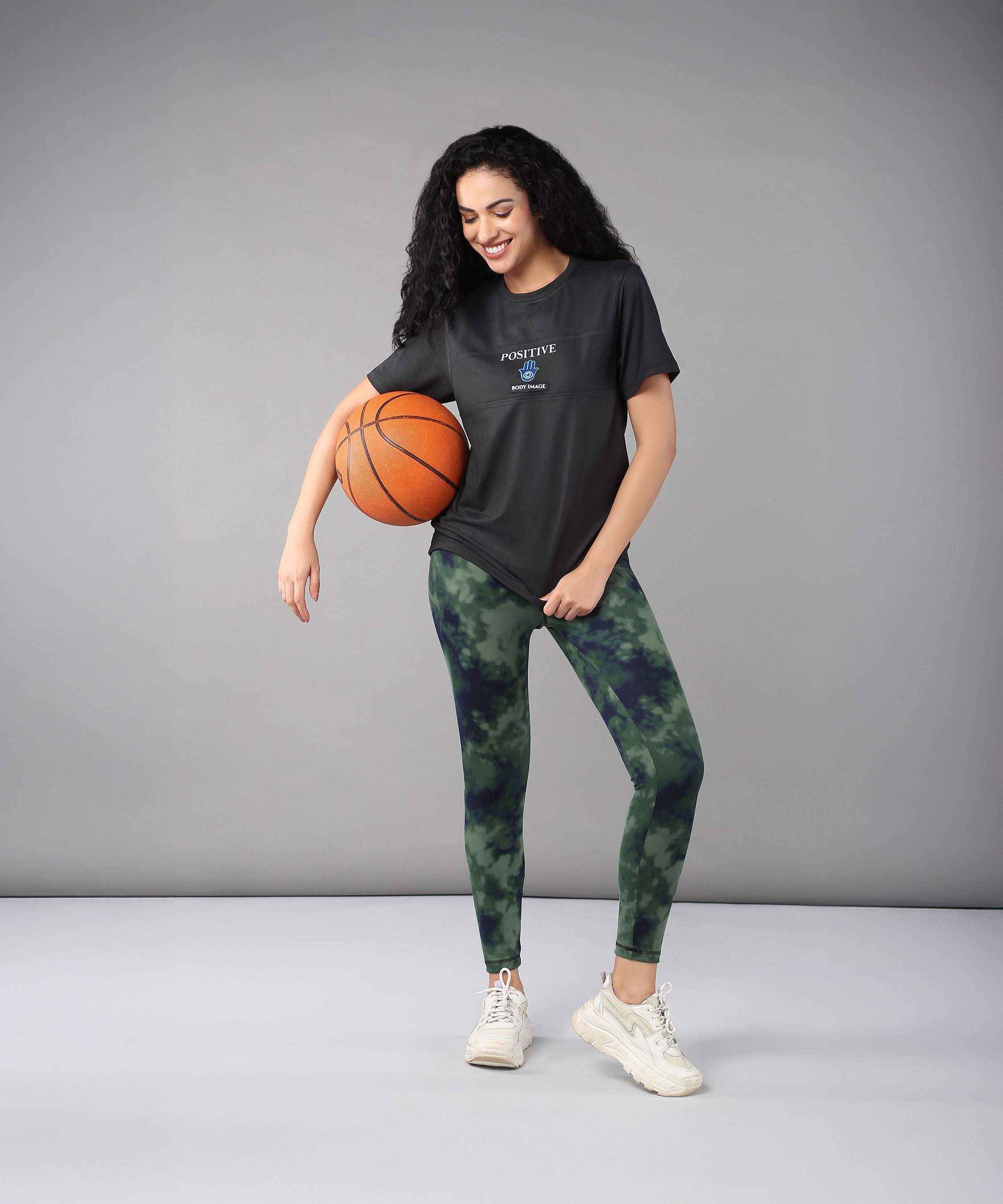 A smiling woman in a Positive Body image T Shirt and sporty outfit holds a basketball against a plain grey background, radiating confidence and positivity.