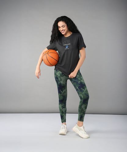A smiling woman in a Positive Body image T Shirt and sporty outfit holds a basketball against a plain grey background, radiating confidence and positivity.