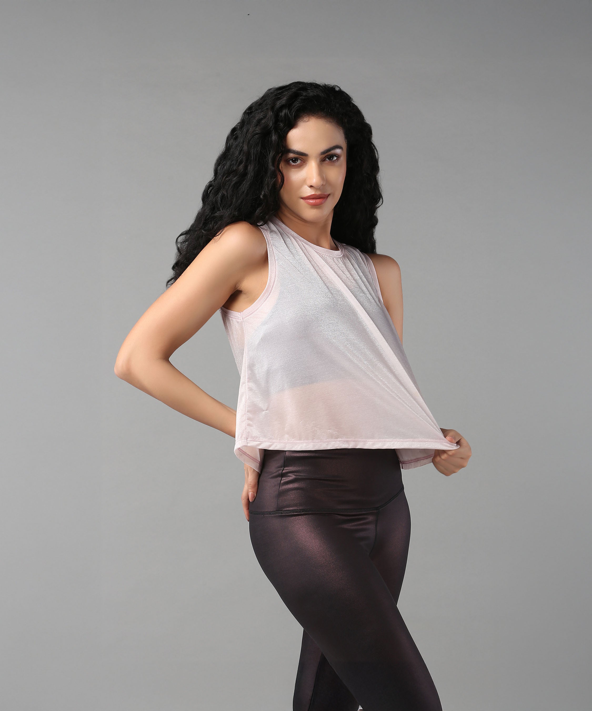 A woman with long curly hair poses against a gray background wearing the Powder Pink Sheer Top and shiny leggings.