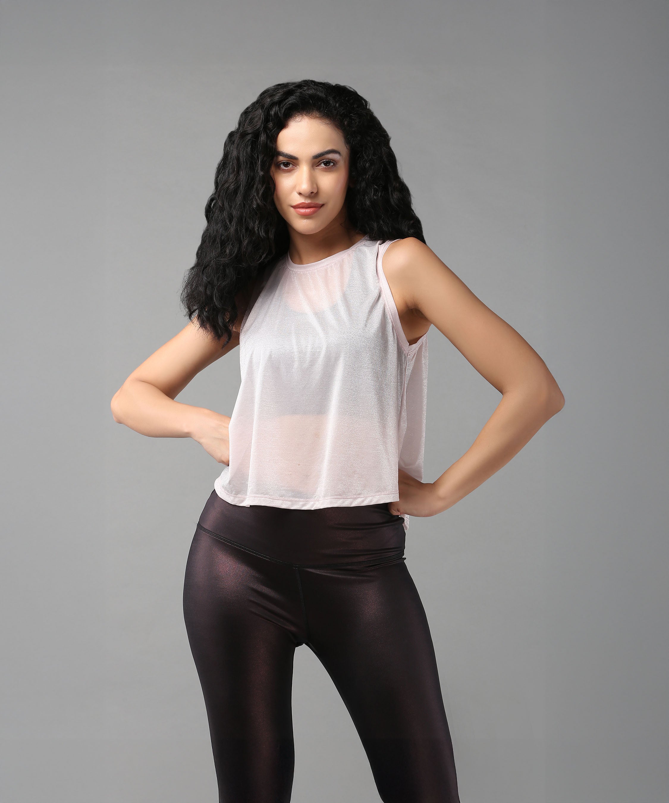 Woman with long curly hair poses confidently in a Powder Pink Sheer Top and black leggings.