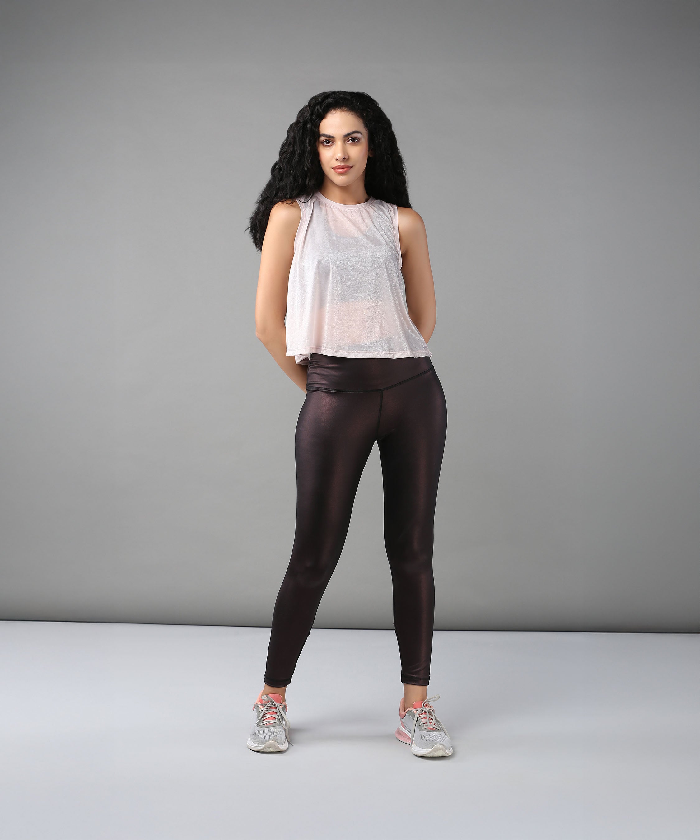 A woman with curly hair models the Powder Pink Sheer Top and shiny black leggings against a plain gray background.