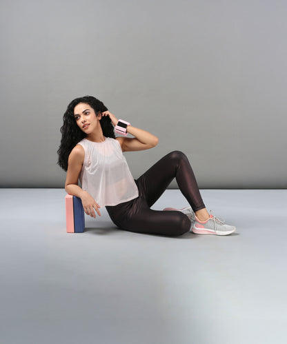A woman in a Powder Pink Sheer Top and workout clothes sits on the floor, leaning against a yoga block and wearing wrist weights.