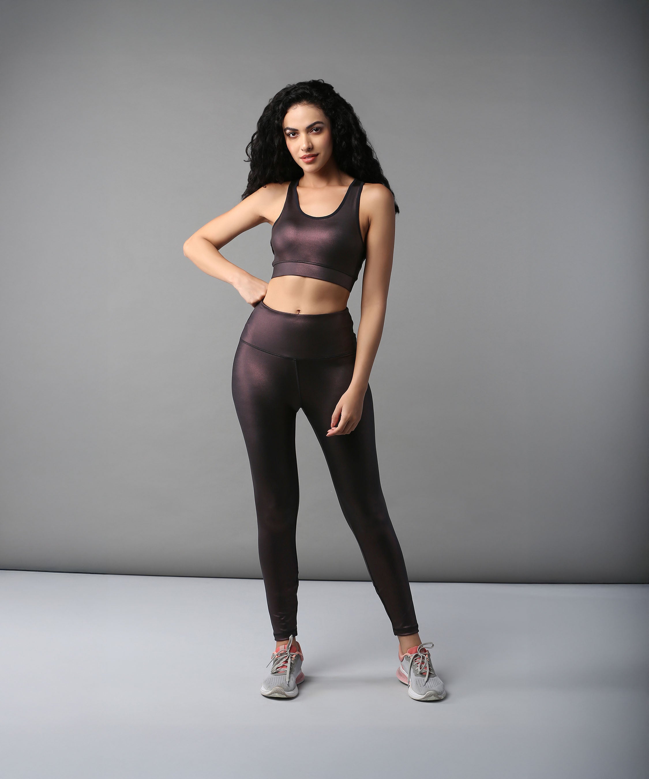 Woman in black activewear and gray sneakers, wearing Radiant Maroon Shimmer Leggings, stands against a plain gray background.