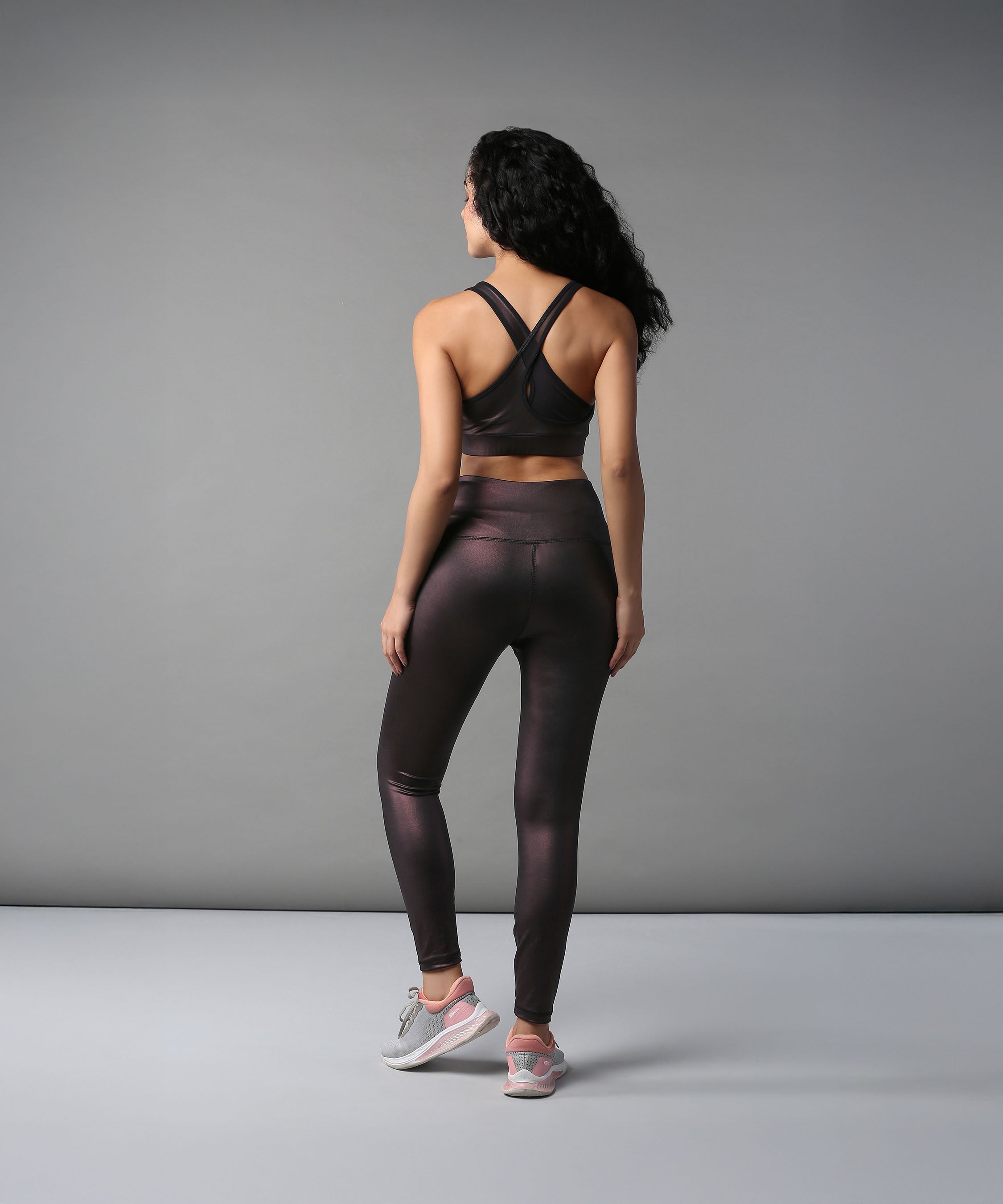 Woman in dark activewear and pink sneakers stands with her back to the camera, showcasing Radiant Maroon Shimmer Leggings against a gray background.