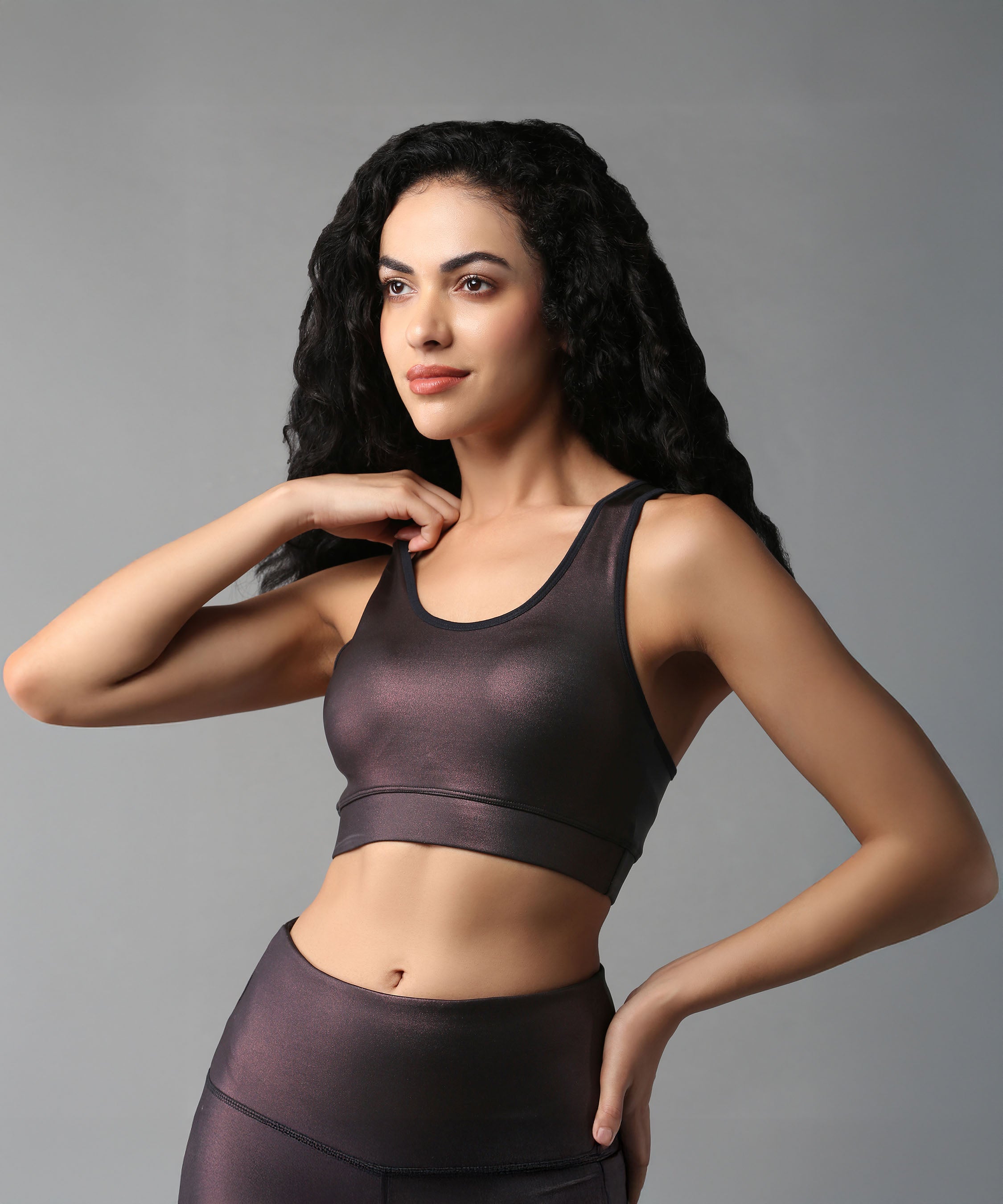 A woman with long curly hair wears the Radiant Maroon Shimmer Sports Bra, posing against a gray background.