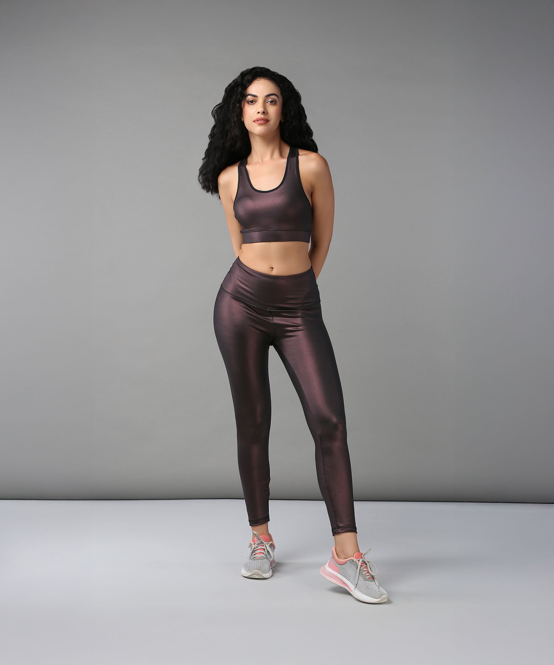 A woman stands confidently in a studio, hands behind her back, wearing the Radiant Maroon Shimmer Sports Bra paired with shiny dark workout clothes.
