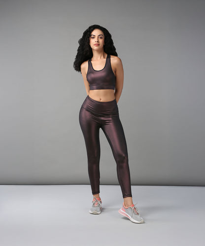 A woman stands confidently in a studio, hands behind her back, wearing the Radiant Maroon Shimmer Sports Bra paired with shiny dark workout clothes.