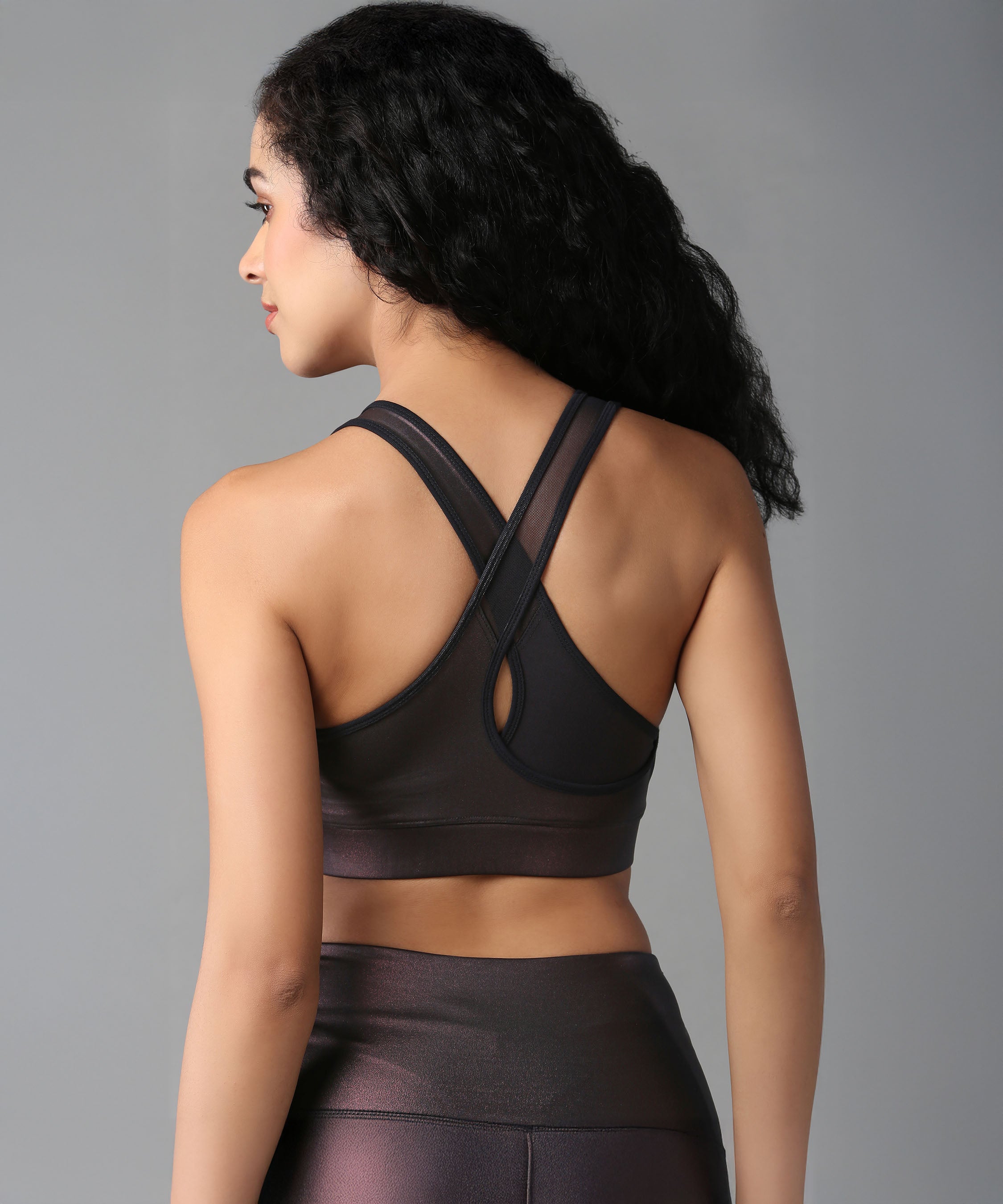 A woman with curly hair is shown from the back against a plain background, wearing the Radiant Maroon Shimmer Sports Bra and matching leggings for high support.