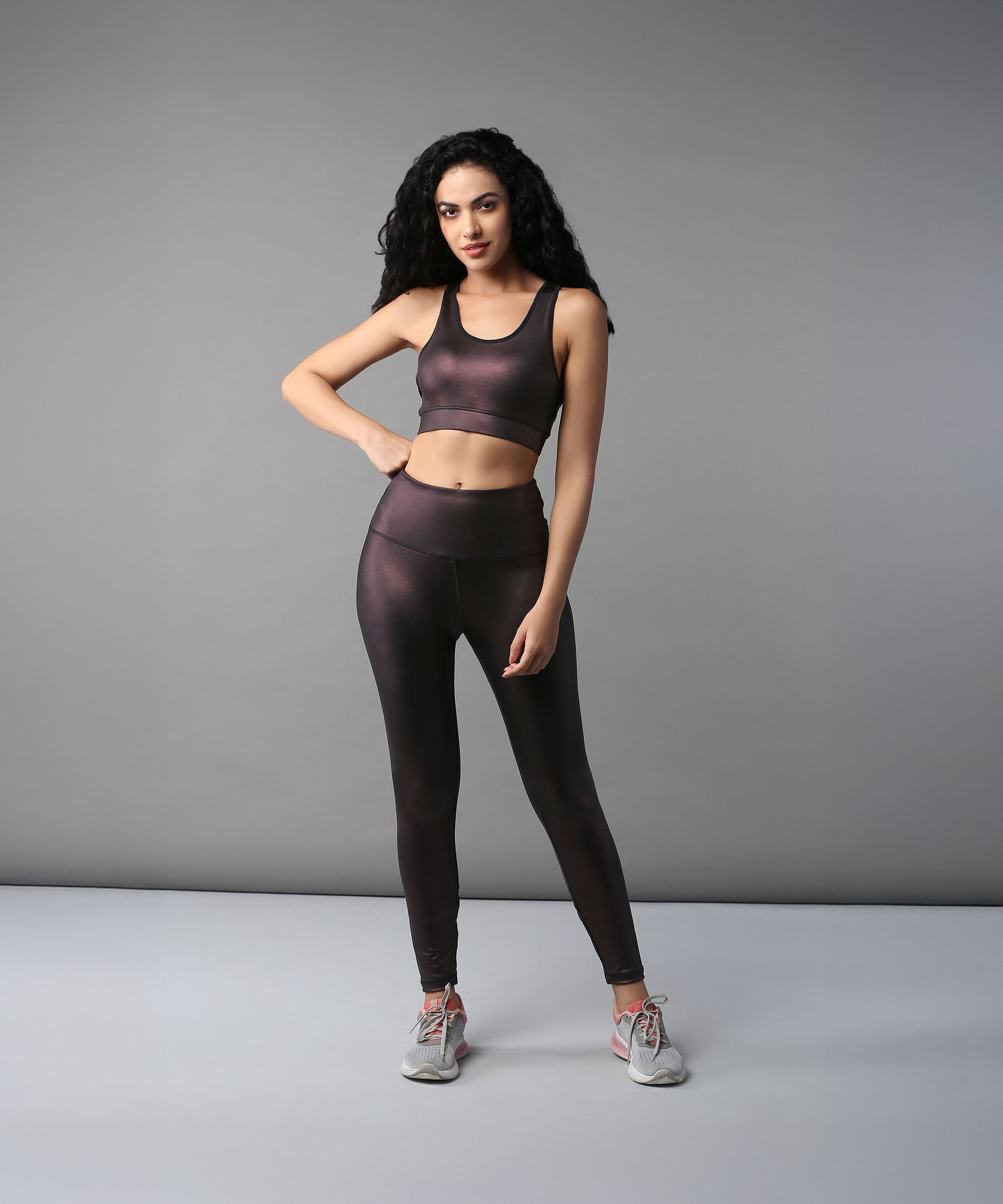 Woman wearing the Radiant Maroon Shimmer Work out Ensemble with cross body racerback and moisture-wicking fabric poses confidently against a plain gray background.