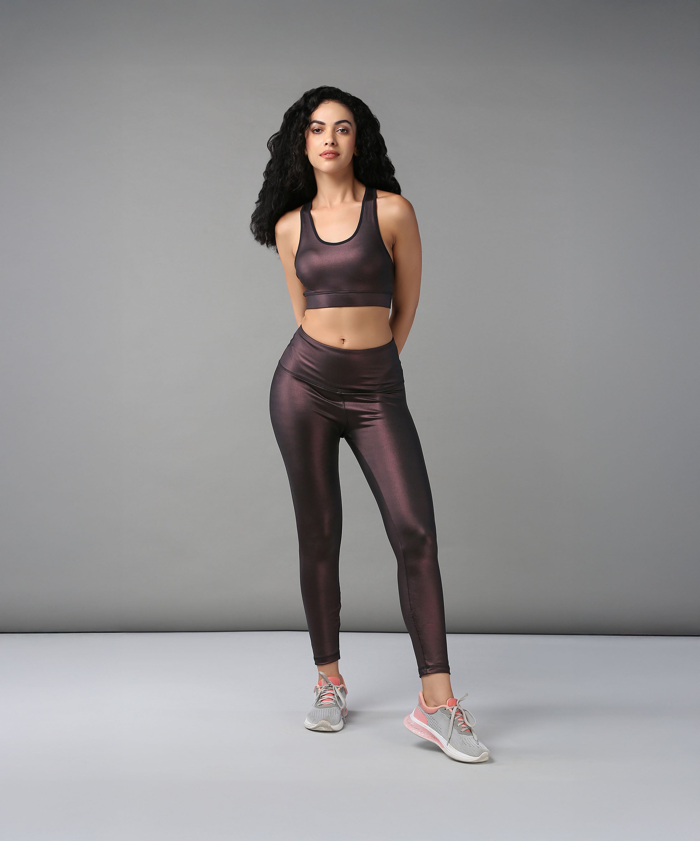 A woman models the Radiant Maroon Shimmer Work out Ensemble—shiny maroon leggings and a racerback crop top—standing in a studio with a grey background, hands behind her back.