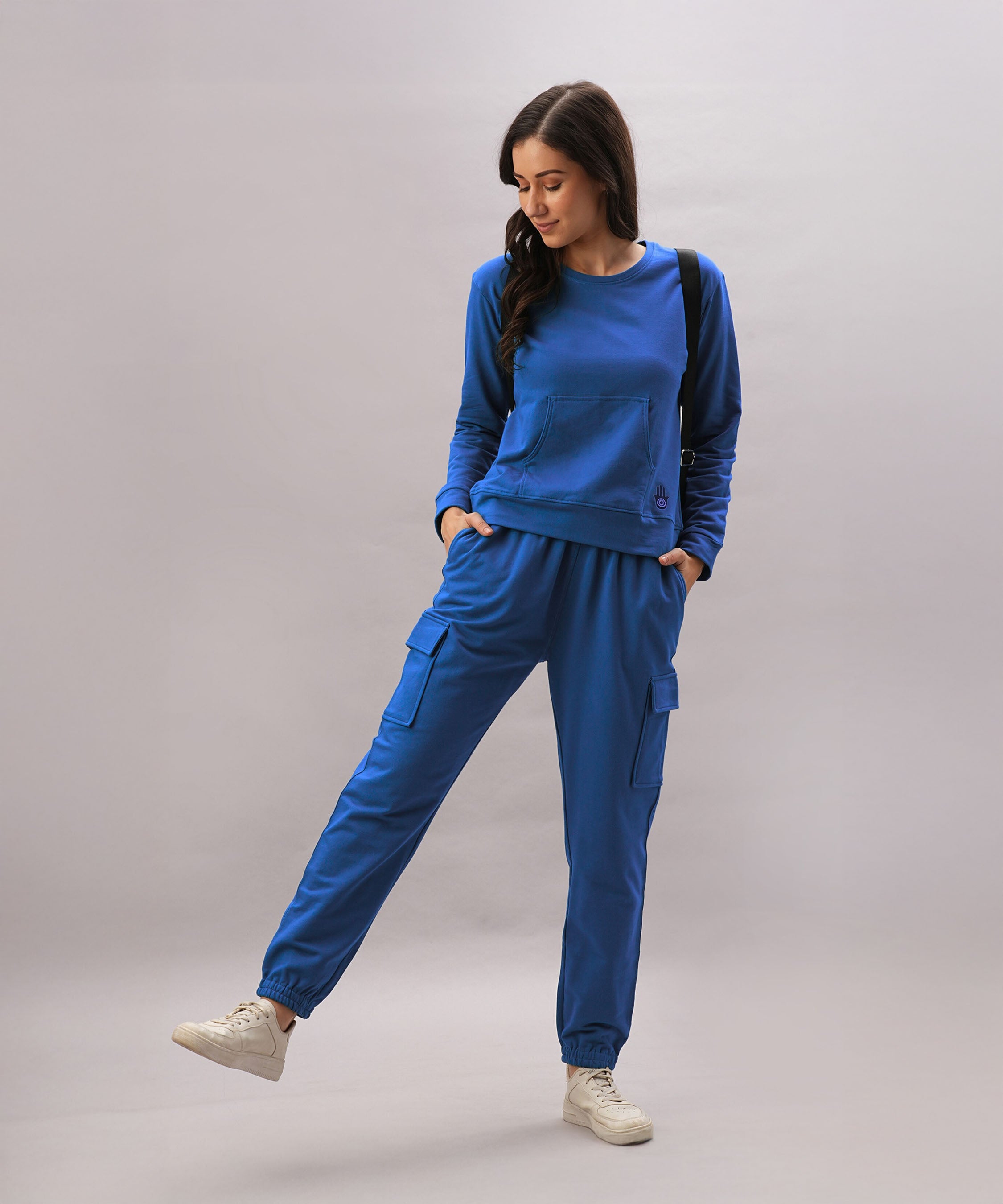 A woman wears the Soft Royal Blue Trackset, featuring cargo-style joggers made from a breathable cotton-viscose blend, paired with white sneakers and a black backpack.