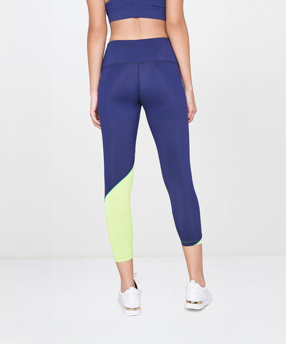 A woman is seen from the back wearing the Lime Tank Top With Leggings, featuring navy leggings with a neon yellow section on one leg, paired with white sneakers and a lime tank top.