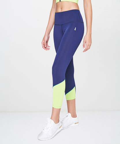 A woman stands confidently against a plain background, wearing Blue and Neon Green Leggings paired with white sneakers and a green sports bra.
