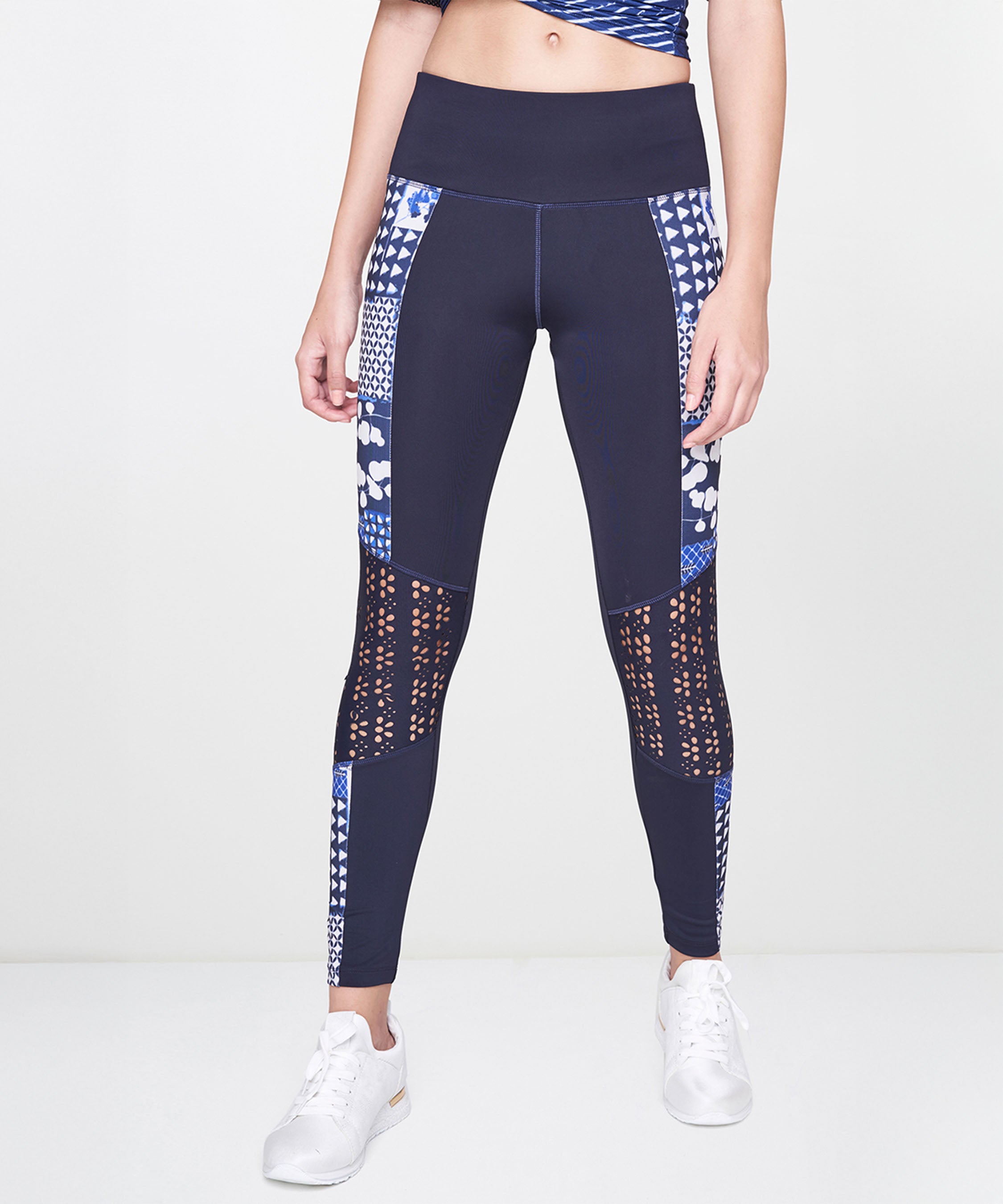 A woman stands against a plain background in Indigo Printed Leggings, featuring a stretchy polyblend fabric and vibrant blue print, paired with white sneakers.