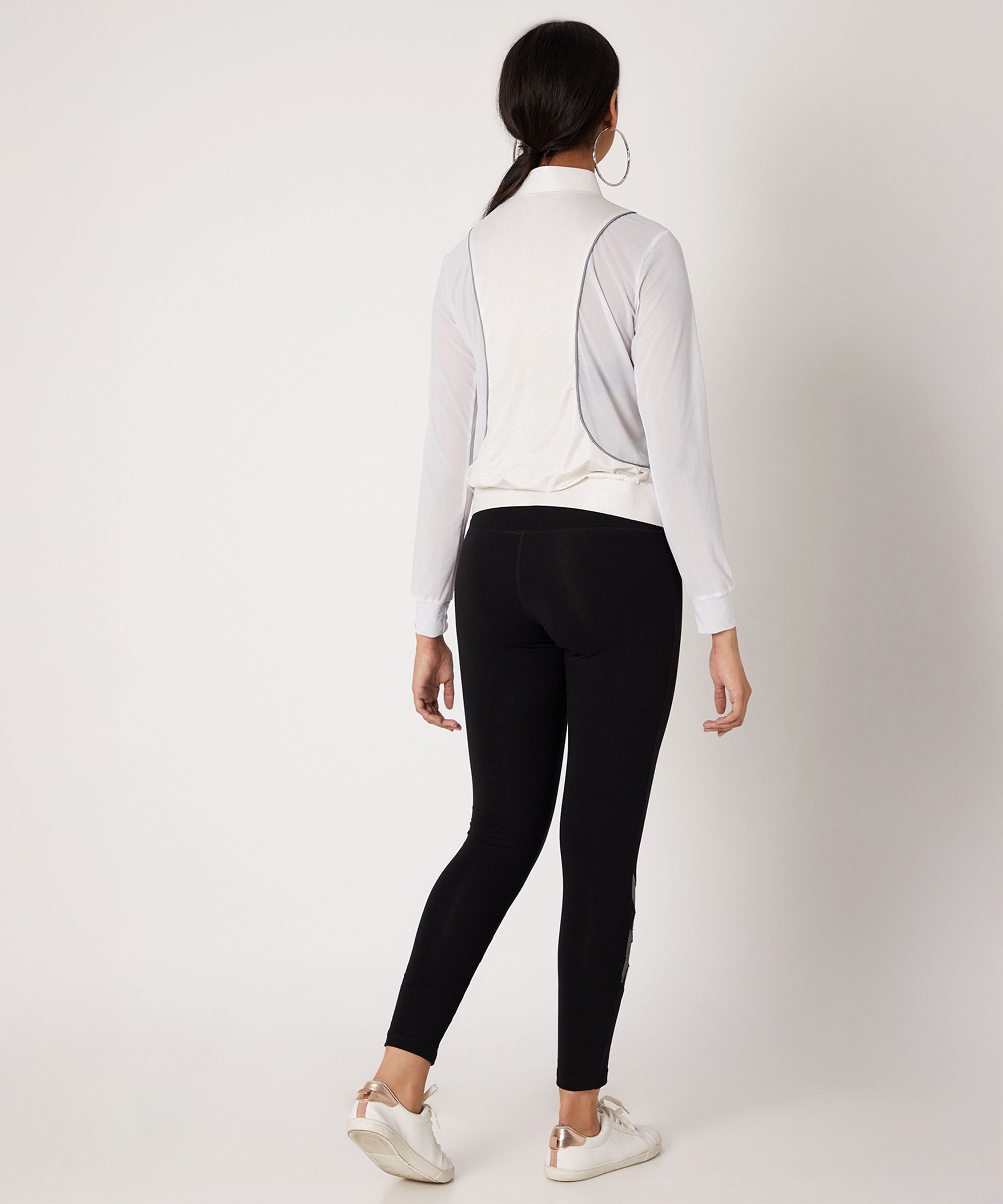 A woman faces away in a white long-sleeve top and Black High Waisted Leggings With a Metallic Touch, made from four-way stretch fabric, paired with white sneakers against a plain background.