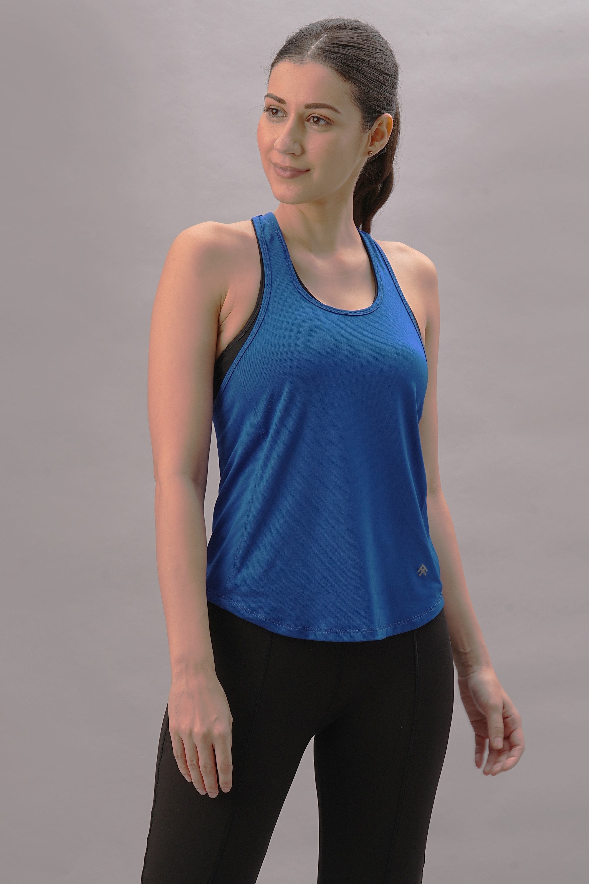 A woman models our Blue Racer Back Top from the activewear collection, paired with black pants, standing against a plain gray background and looking to the side for a sporty, stylish look.
