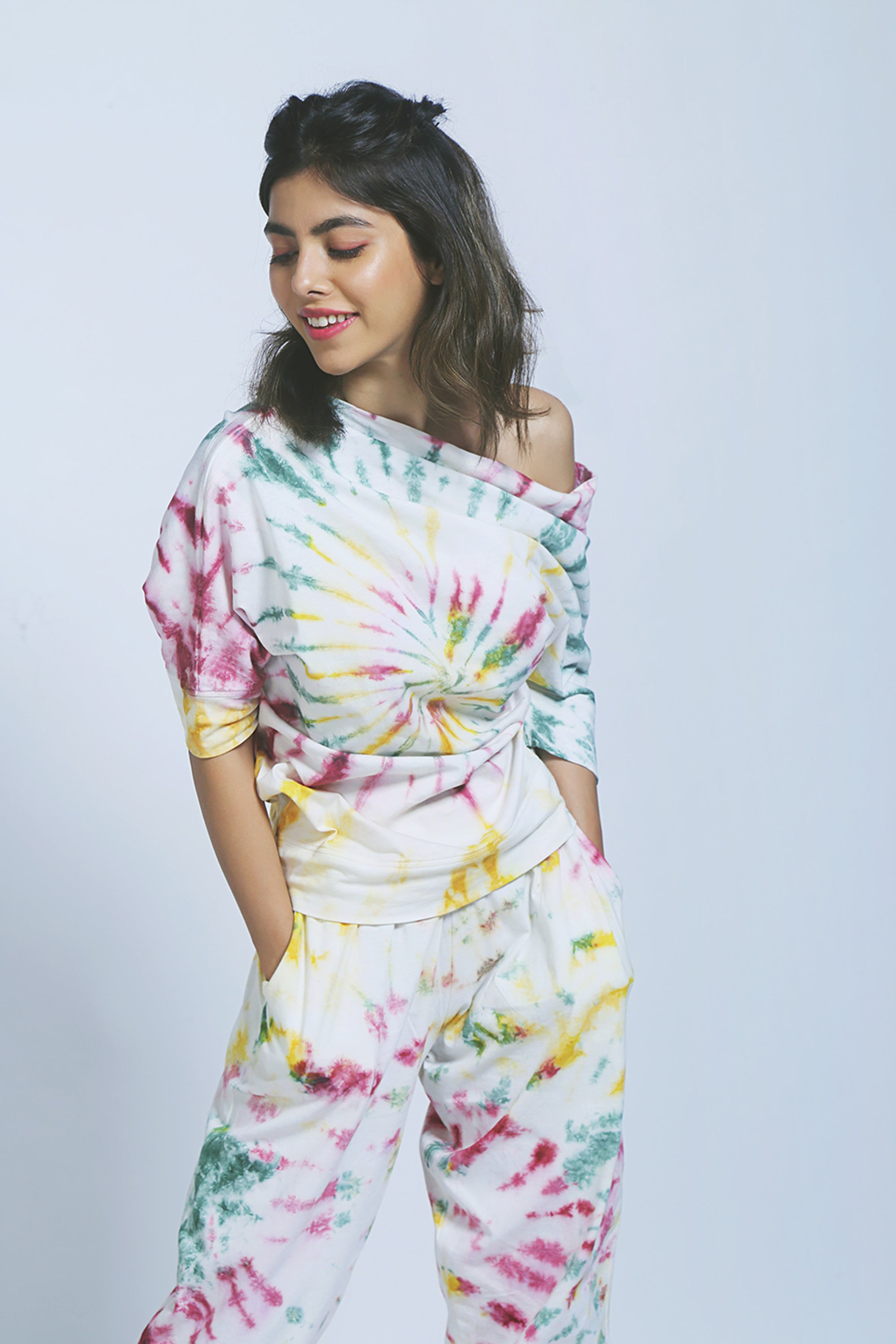 Smiling woman wearing the One Side Off-Shoulder top and matching yoga pants in colorful tie-dye organic cotton, with hands in pockets.