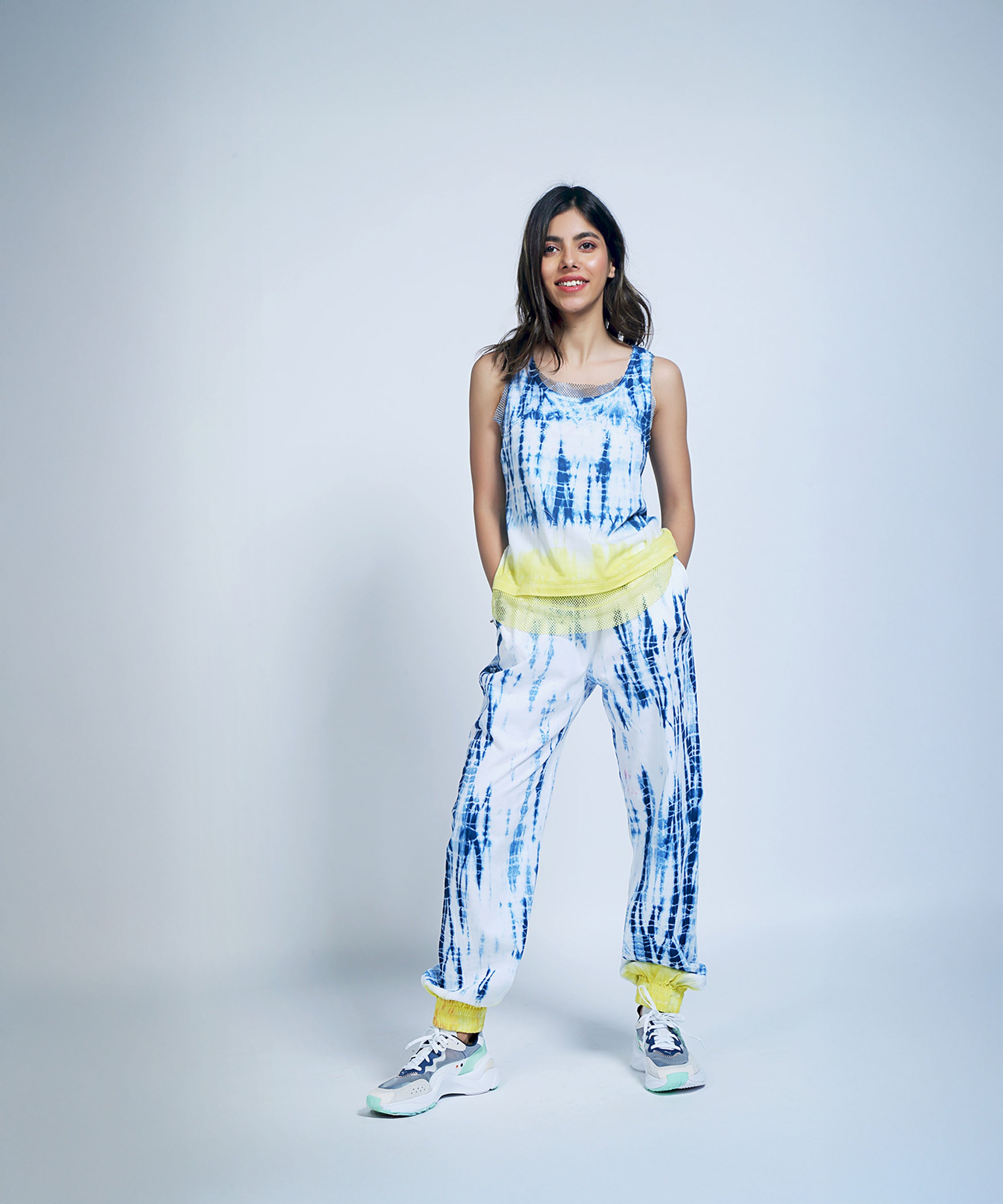 Smiling woman stands with hands in pockets, wearing Comfort Fit Joggers + Netted Tank Top in blue and white tie-dye with yellow accents. Made from organic cotton, these joggers offer a relaxed vibe for any occasion.