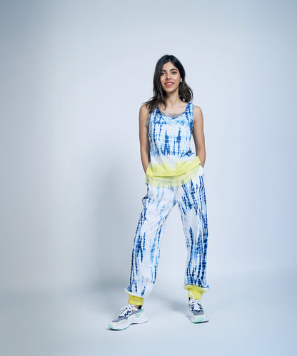 Smiling woman stands with hands in pockets, wearing Comfort Fit Joggers + Netted Tank Top in blue and white tie-dye with yellow accents. Made from organic cotton, these joggers offer a relaxed vibe for any occasion.