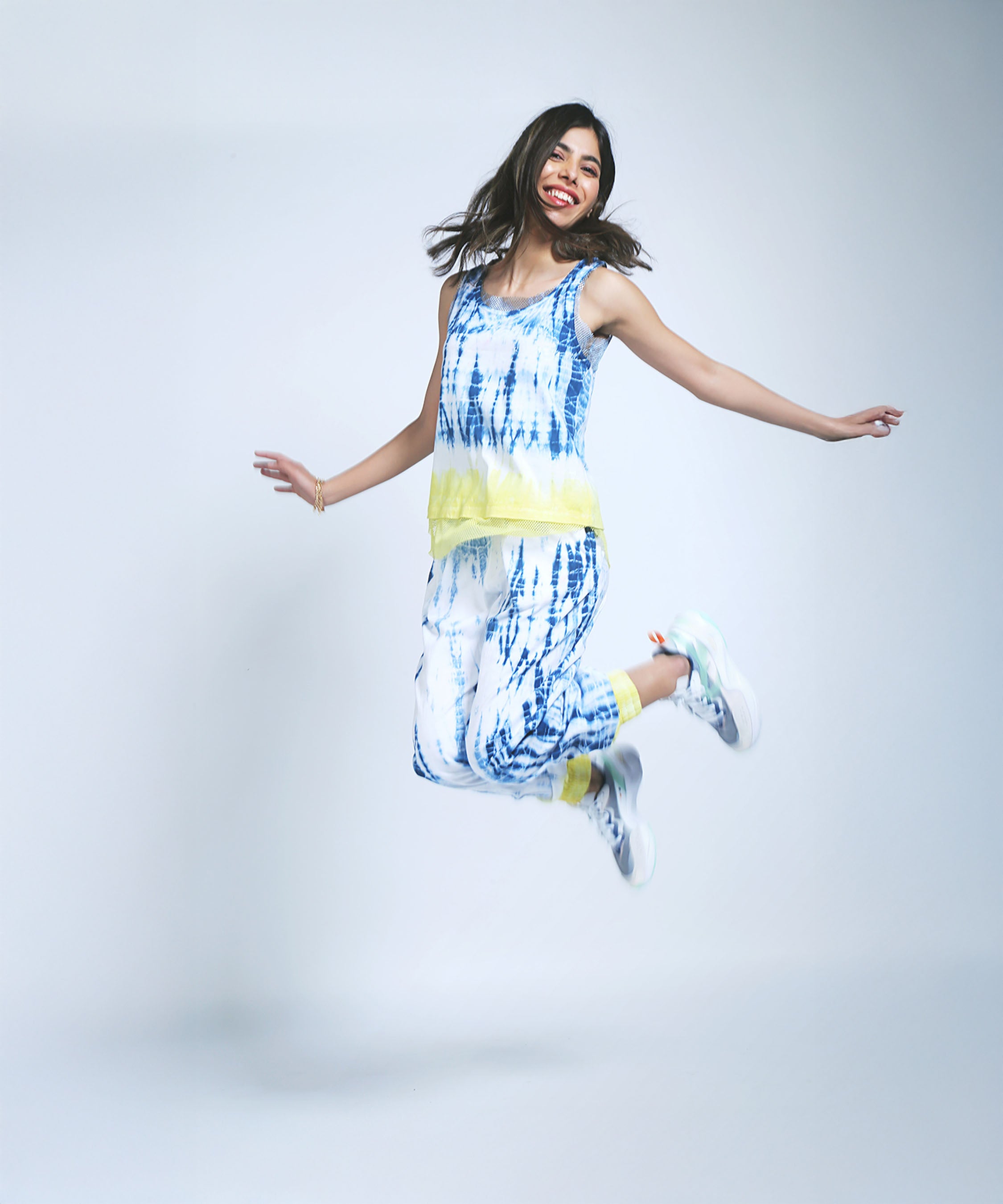 A smiling woman jumps joyfully against a plain light background, wearing Comfort Fit Joggers and a Netted Tank Top in blue and yellow tie-dye made with eco-friendly dyes.